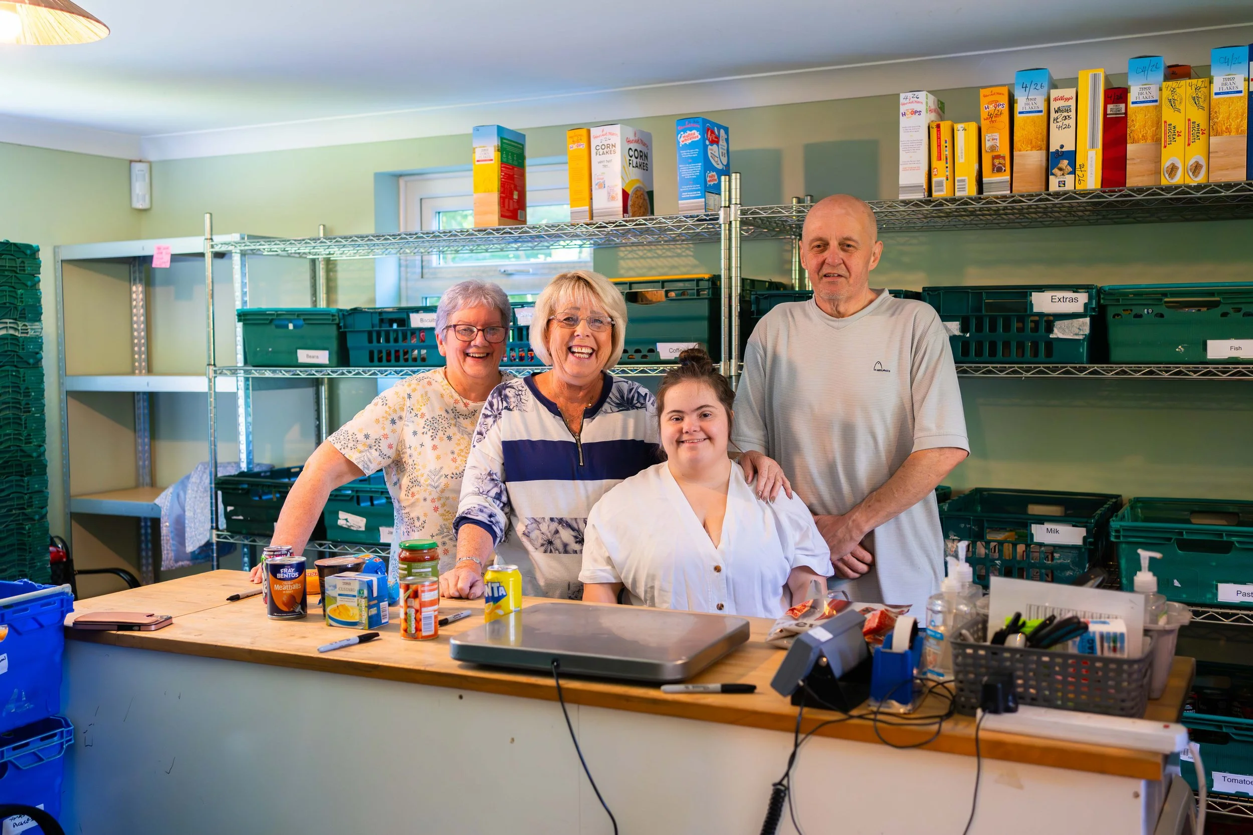 Four people standing behind a counter in a food bank or community kitchen, with canned goods, food packages, and a scale on the counter, and shelves with labeled green bins and cereal boxes in the background.