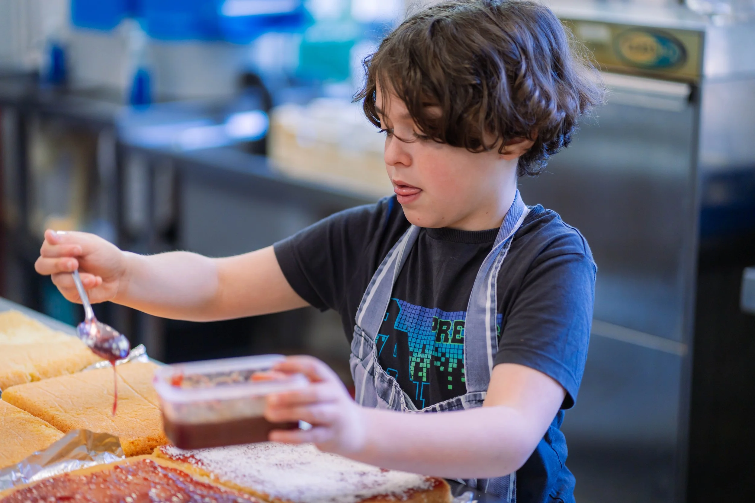 A young boy with curly brown hair and a gray apron is spreading chocolate sauce onto a cake with a spoon in a kitchen.