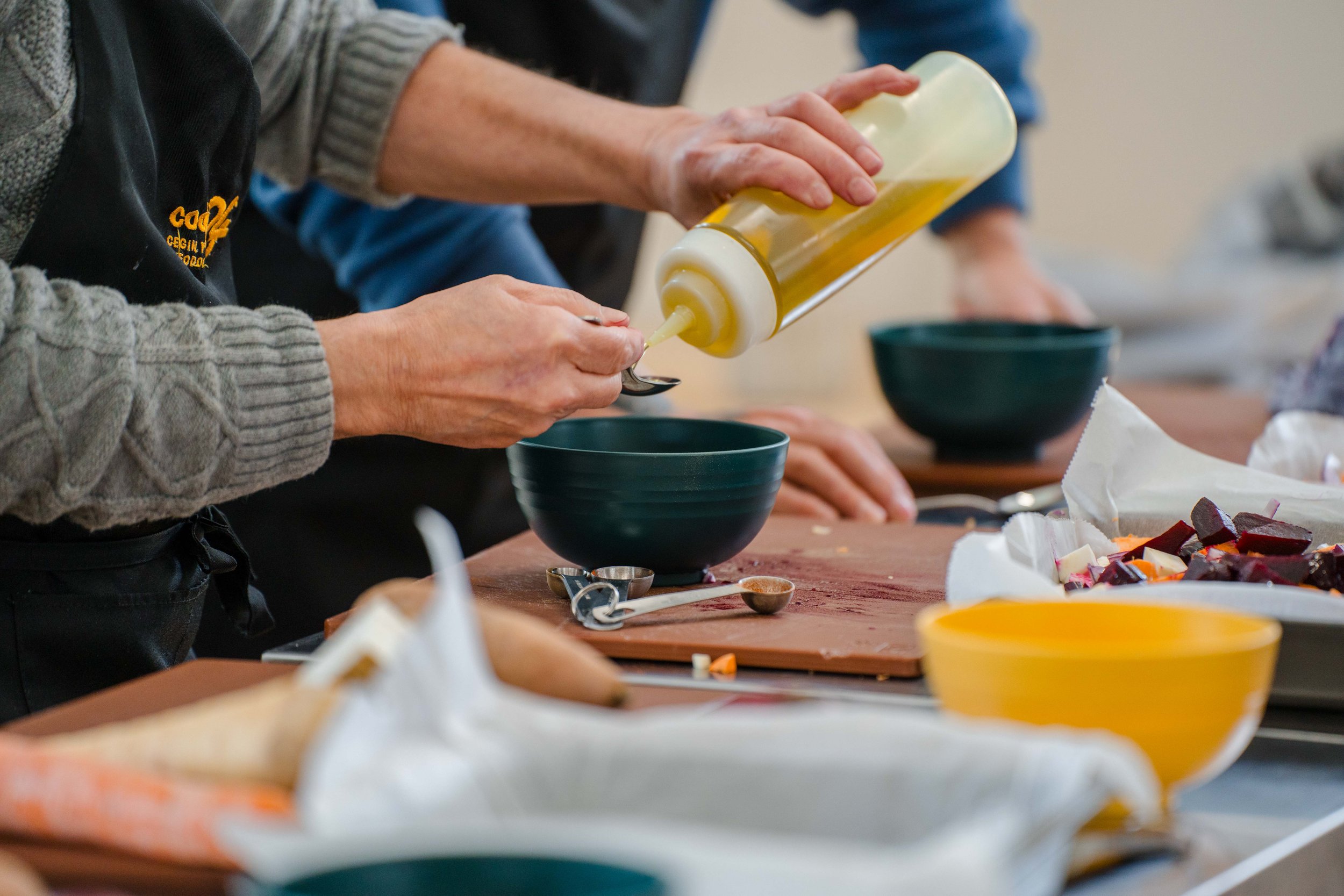 A person pouring mayonnaise into a green bowl at a kitchen counter, with various bowls, spoons, and chopped vegetables nearby.