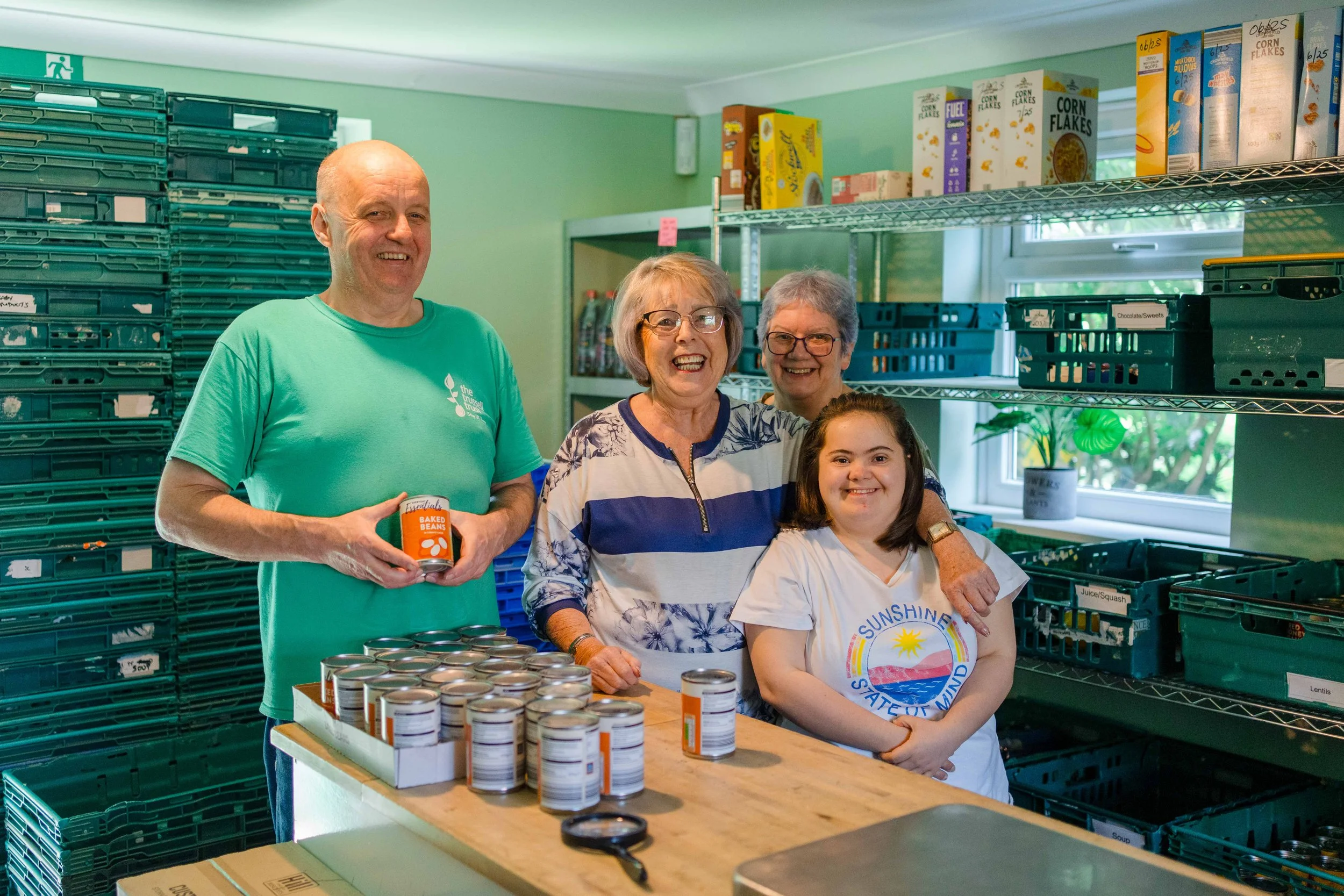 Four smiling people standing in a food pantry with stacked canned goods and shelves of cereal and supplies behind them.
