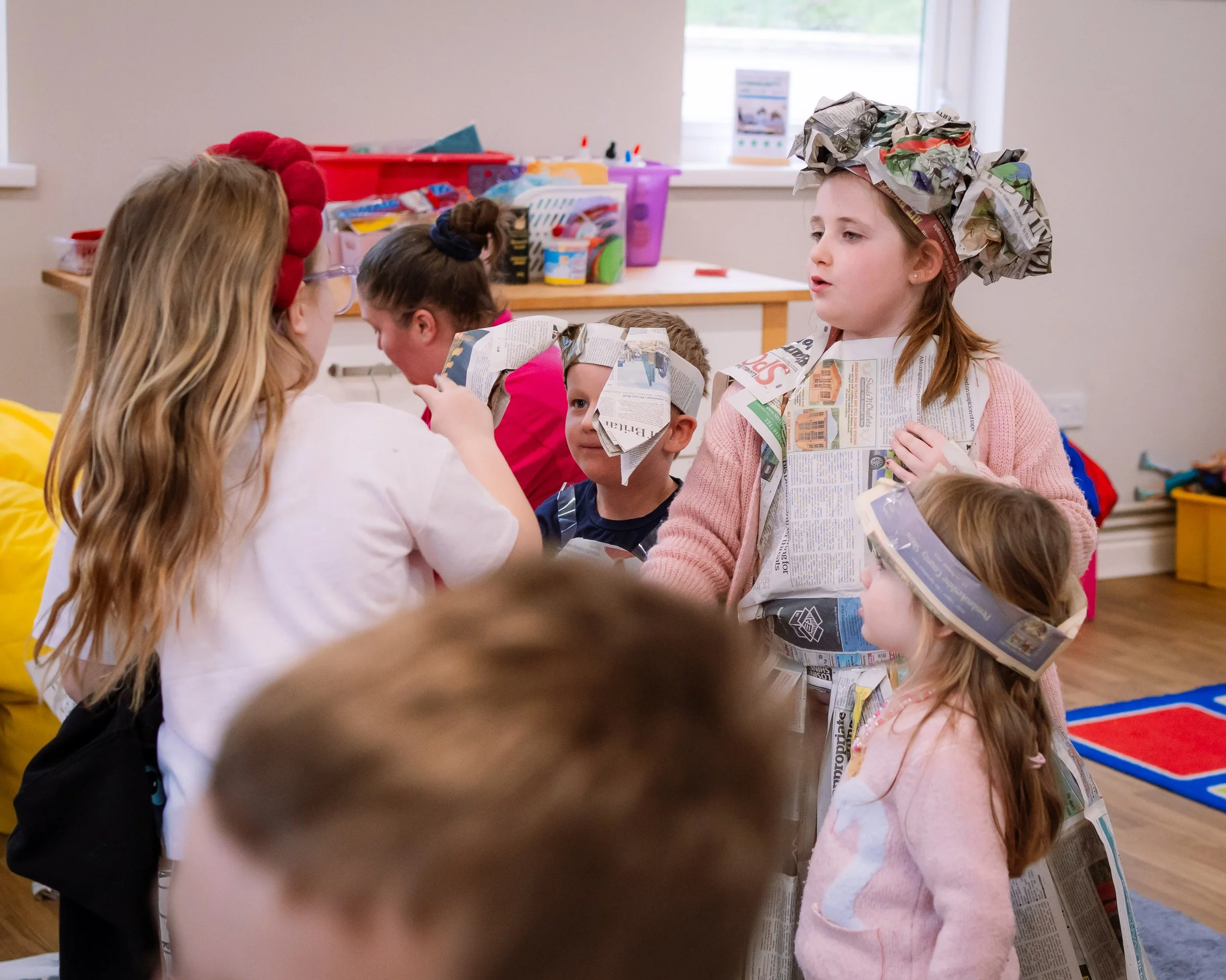 Children participating in a classroom activity wearing newspaper costumes and hats, engaging in a group discussion.