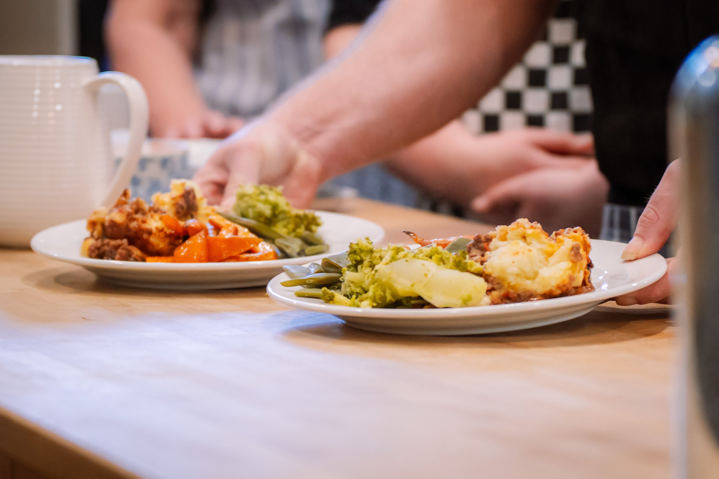 People serving plates with mashed potatoes, broccoli, green beans, carrots, and meatloaf, on a wooden table.