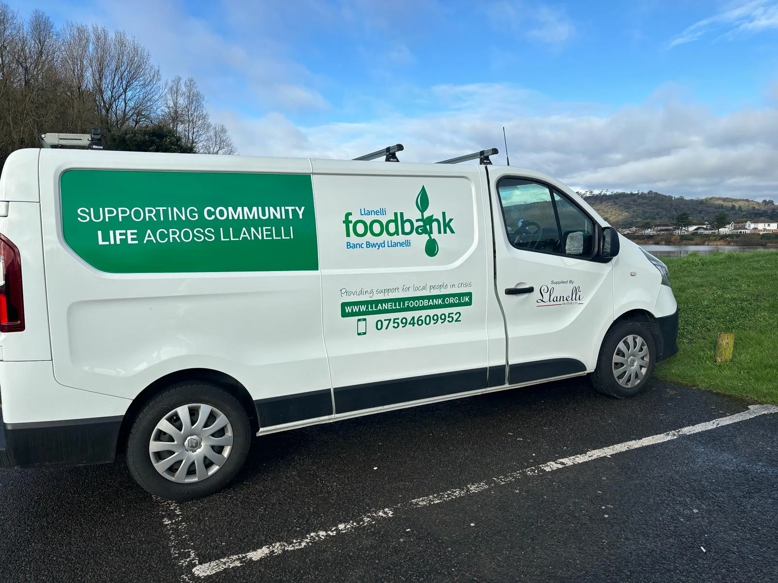 White van with green signage supporting community foodbank services, parked beside a grassy area near rivers and trees.