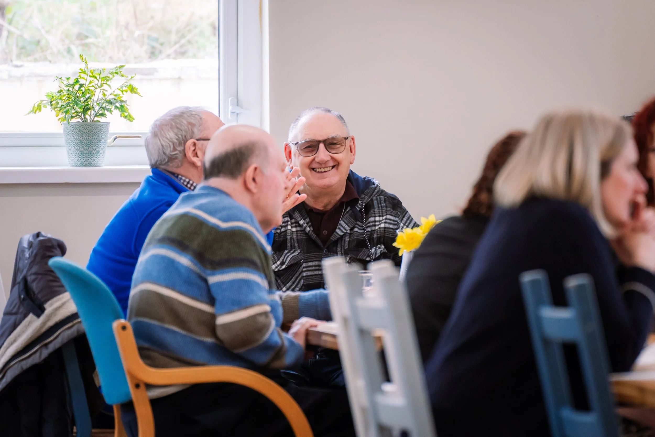 A group of older adults sitting around a table, smiling and engaging with each other during a gathering or meeting. There are potted plants on the windowsill and a vase of yellow flowers on the table.