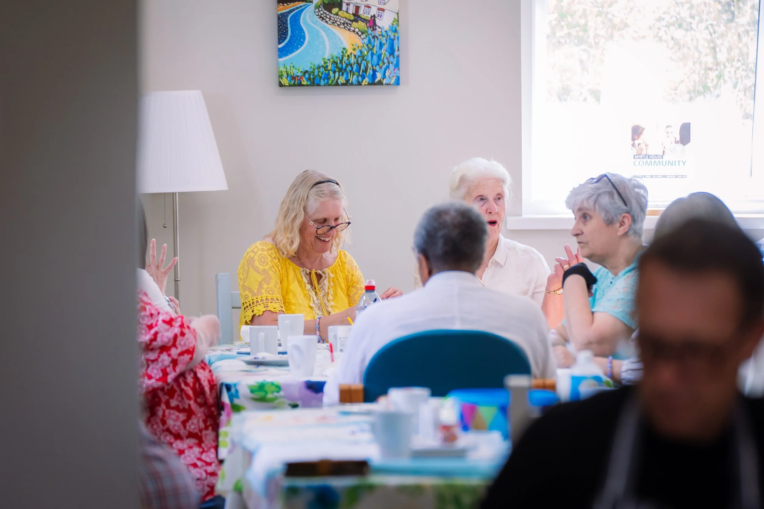 Group of elderly women and men sitting at a table, engaging in conversation, in a bright room with a colorful painting on the wall and an open window.