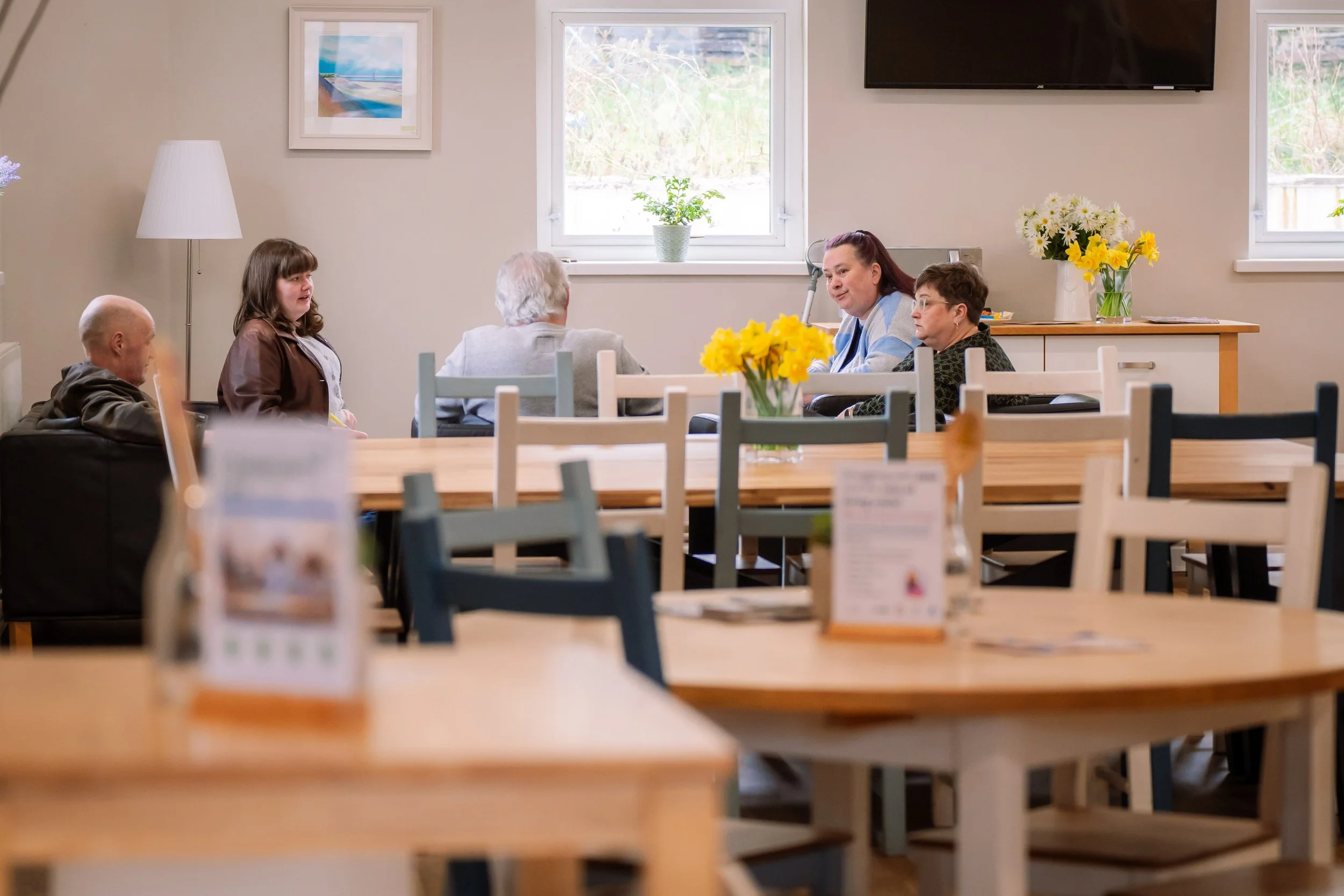 Interior of a dining area with four elderly women, a teenage girl, and a staff member engaged in conversation. Bright and welcoming with floral decor and several tables and chairs.