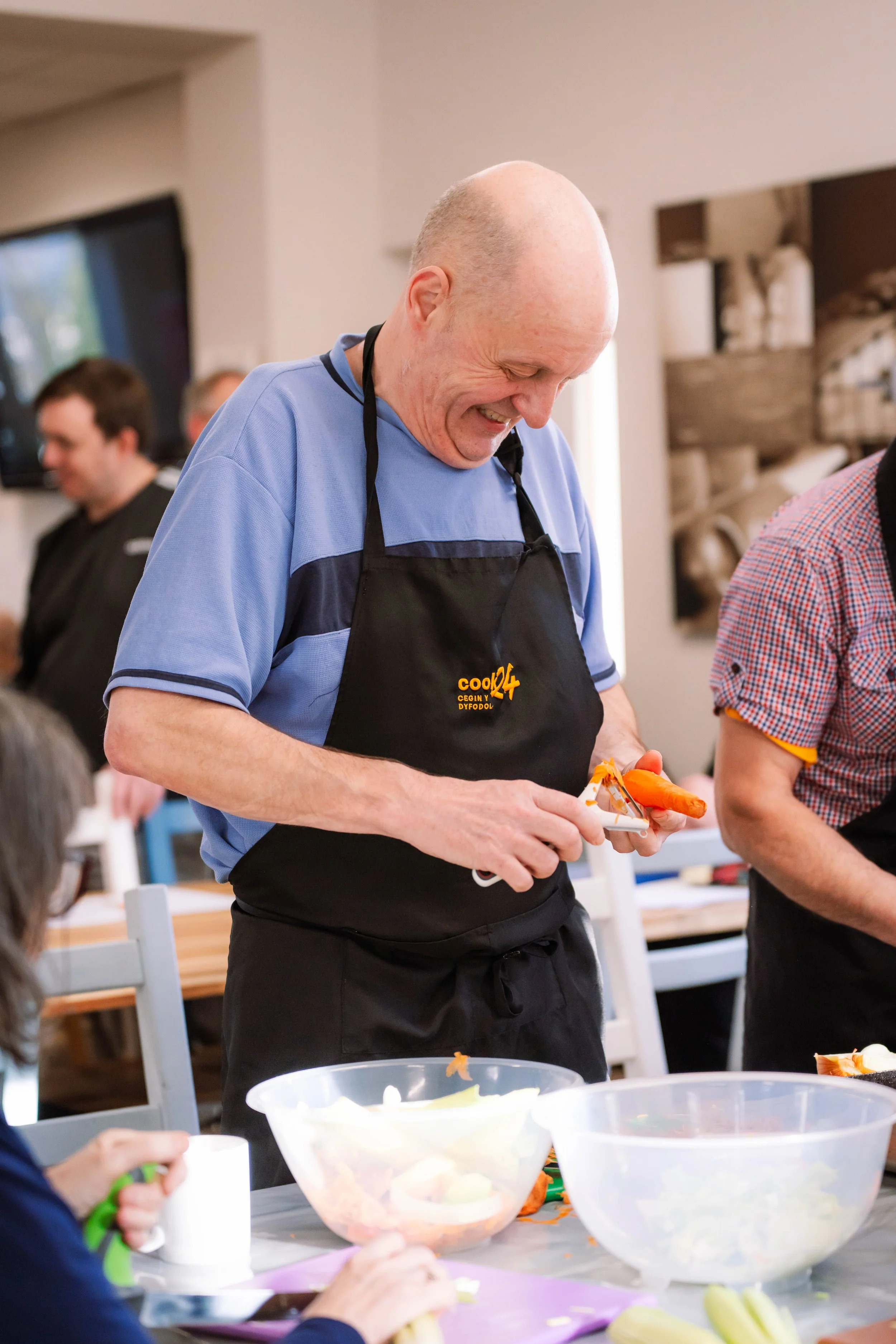 A man wearing a black apron with yellow text and a blue shirt, smiling and preparing food at a cooking class or gathering.
