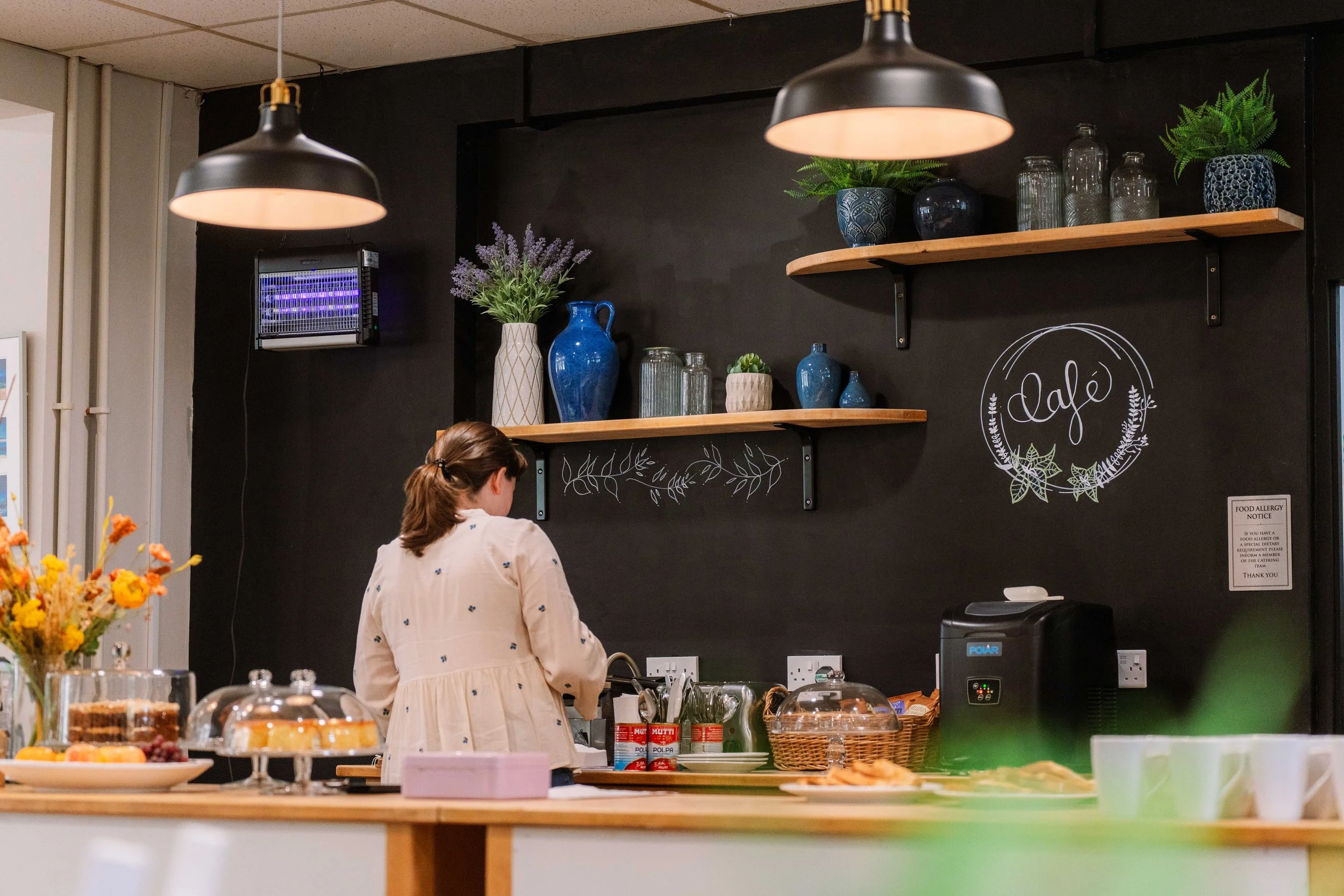 A woman with brown hair in a ponytail, wearing a cream-colored dress, stands behind a counter in a cozy cafe or bakery. The counter displays cakes, pastries, and flowers. The background features a black chalkboard wall with handwritten white chalk art that says 'Café' and decorated with hanging shelves holding vases, jars, and plants. Two black pendant lights hang overhead.