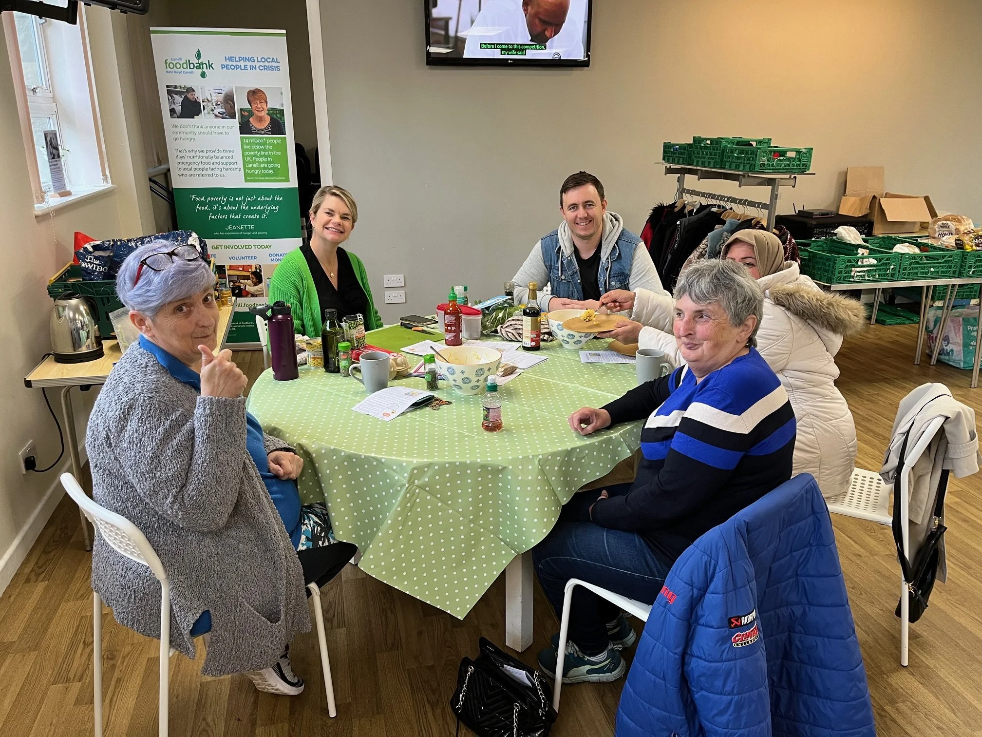 A group of five people sitting around a table with a green polka dot tablecloth, enjoying a meal with condiments and drinks. They are indoors, in a room with a banner for Foodbank in the background and packing supplies around.