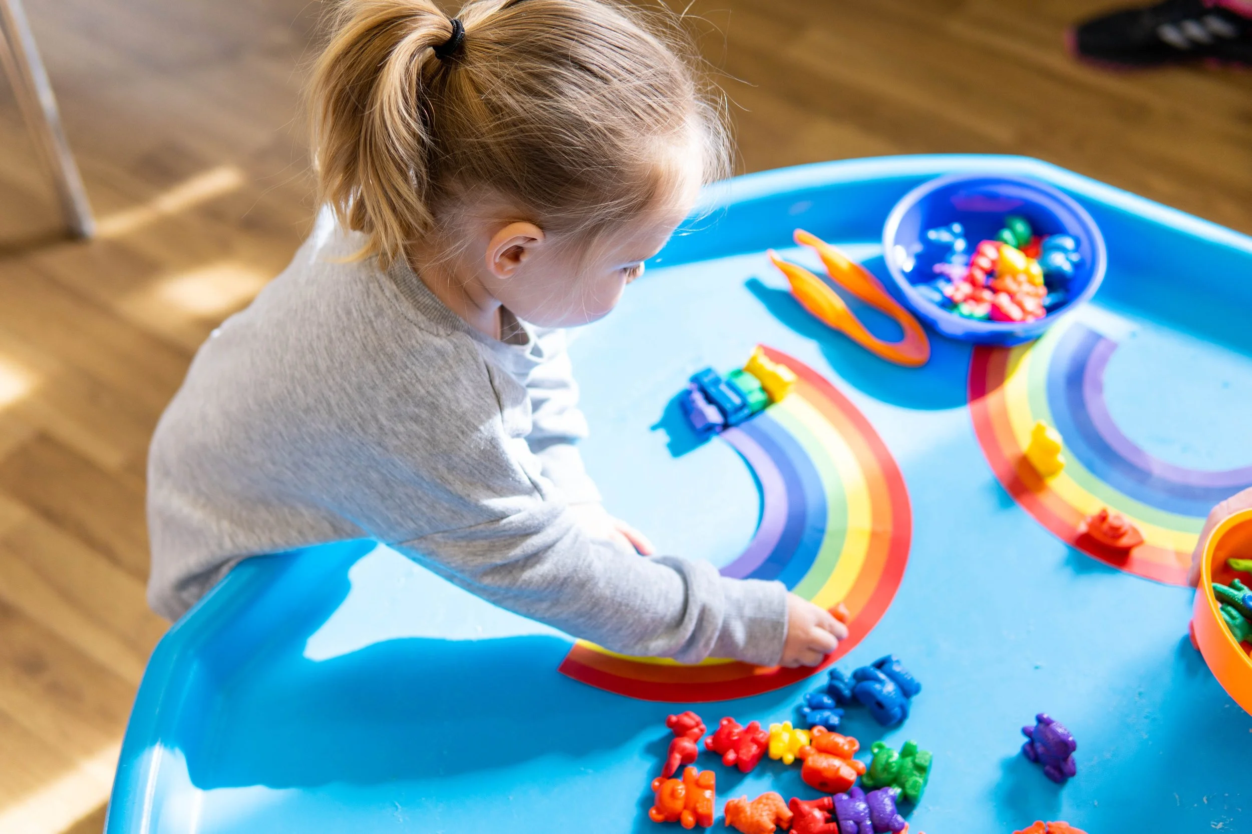 A young girl with a ponytail is playing with rainbow-colored paper strips and small rainbow teddy bear toys on a blue table. There are also two bowls with small toy figures and a pair of orange scissors on the table.
