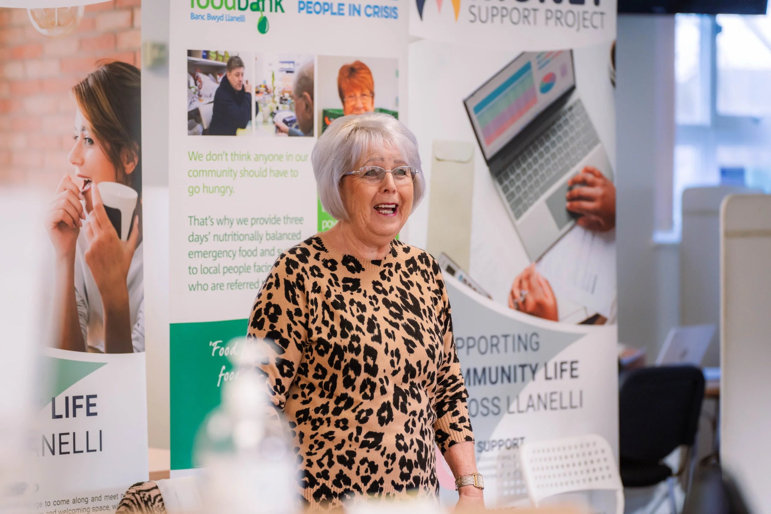 An elderly woman with gray hair and glasses, wearing a leopard print top, smiling and standing in front of a community support booth with informational posters.