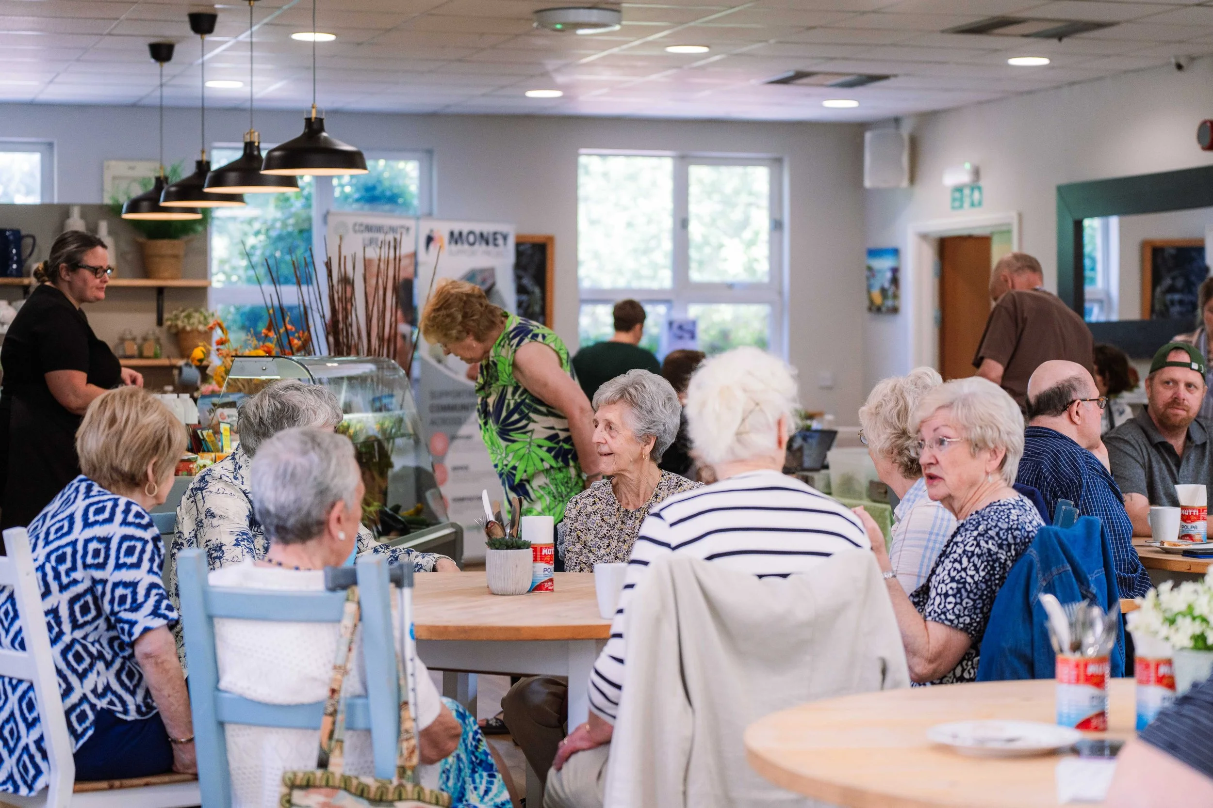 Elderly people seated around tables in a community dining hall, engaging in conversation and enjoying a meal. There are staff members behind a counter in the background, with sunlight coming through the windows.