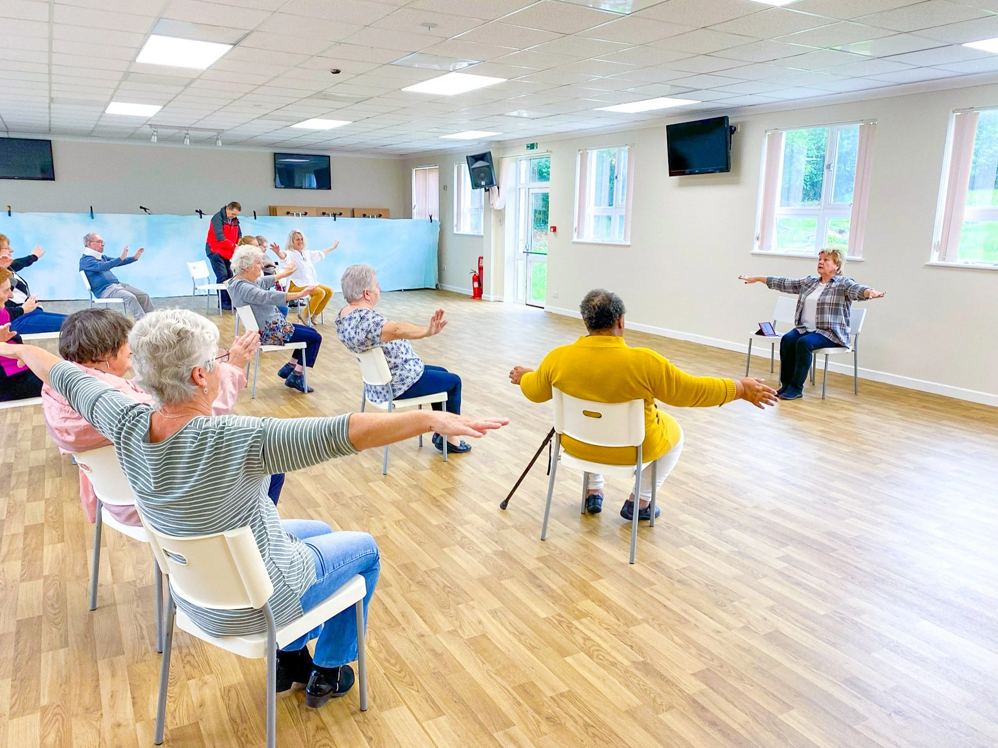 Group of elderly people participating in seated exercise class led by instructor in spacious room with wooden floor and large windows.