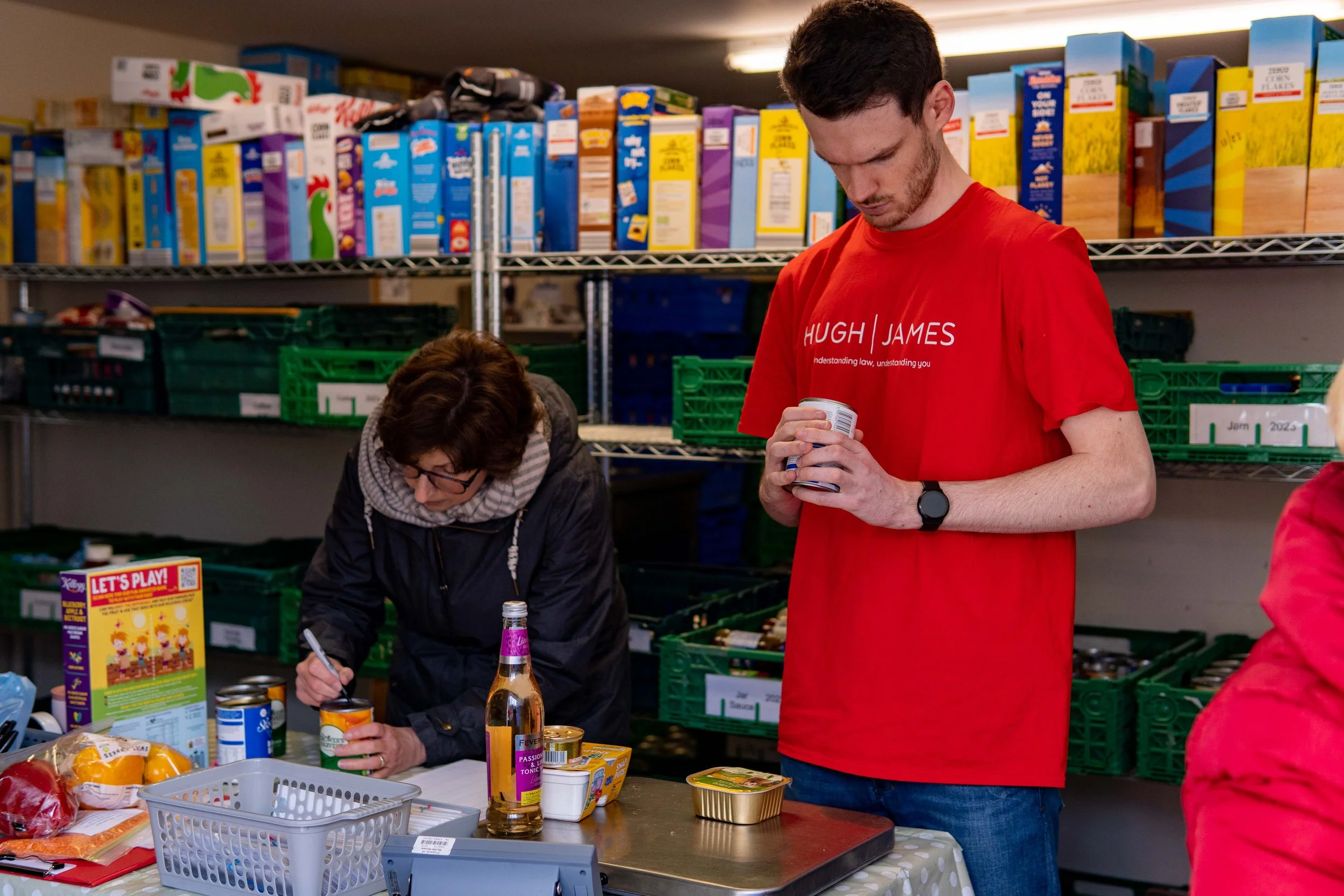 A man in a red shirt with white text standing and holding a can, a woman signing a can while working at a table, in a storage room with shelves of food items.