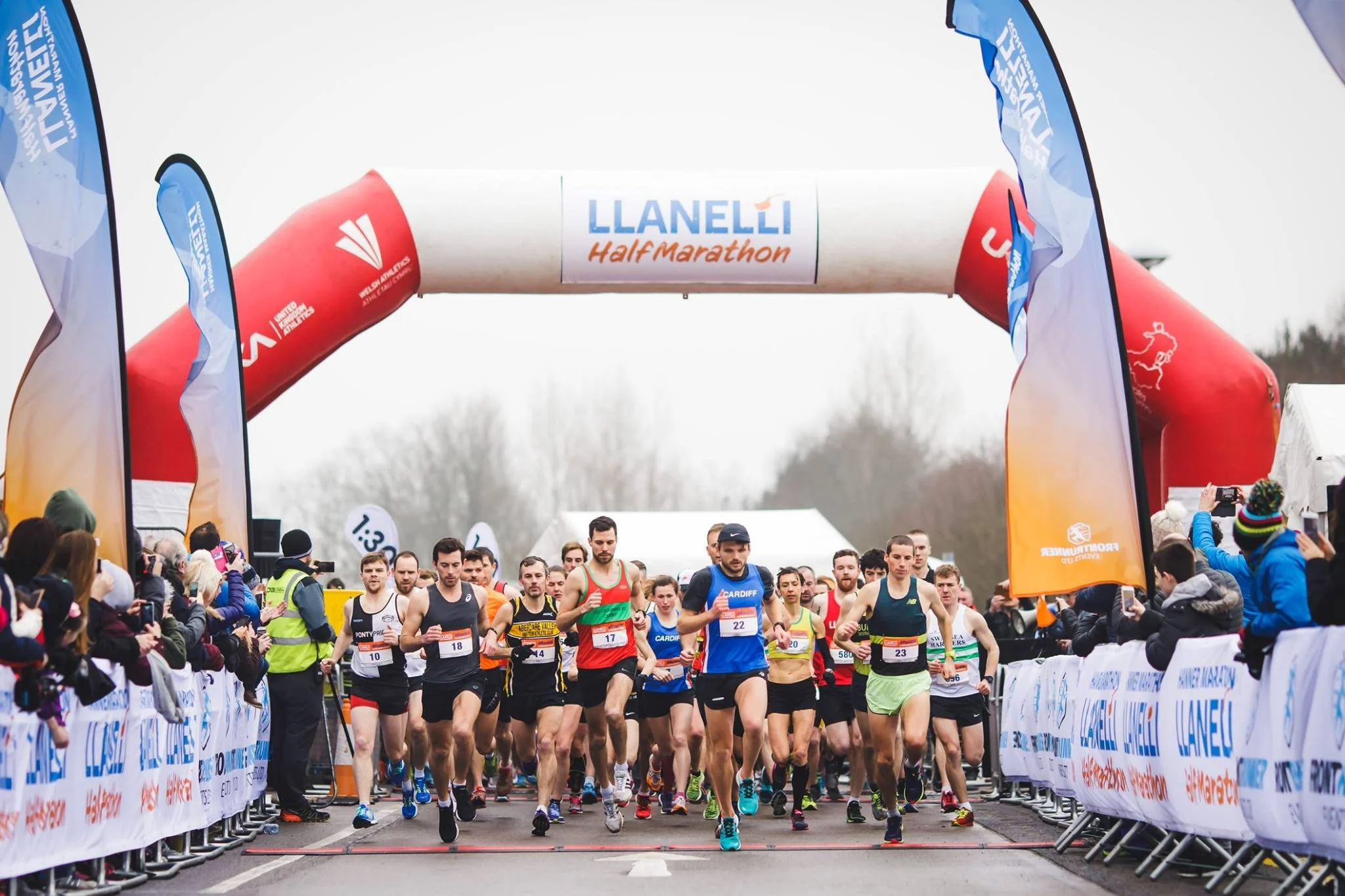 Runners at the start of a half marathon race under an inflatable archway with a crowd on both sides taking photos and cheering.