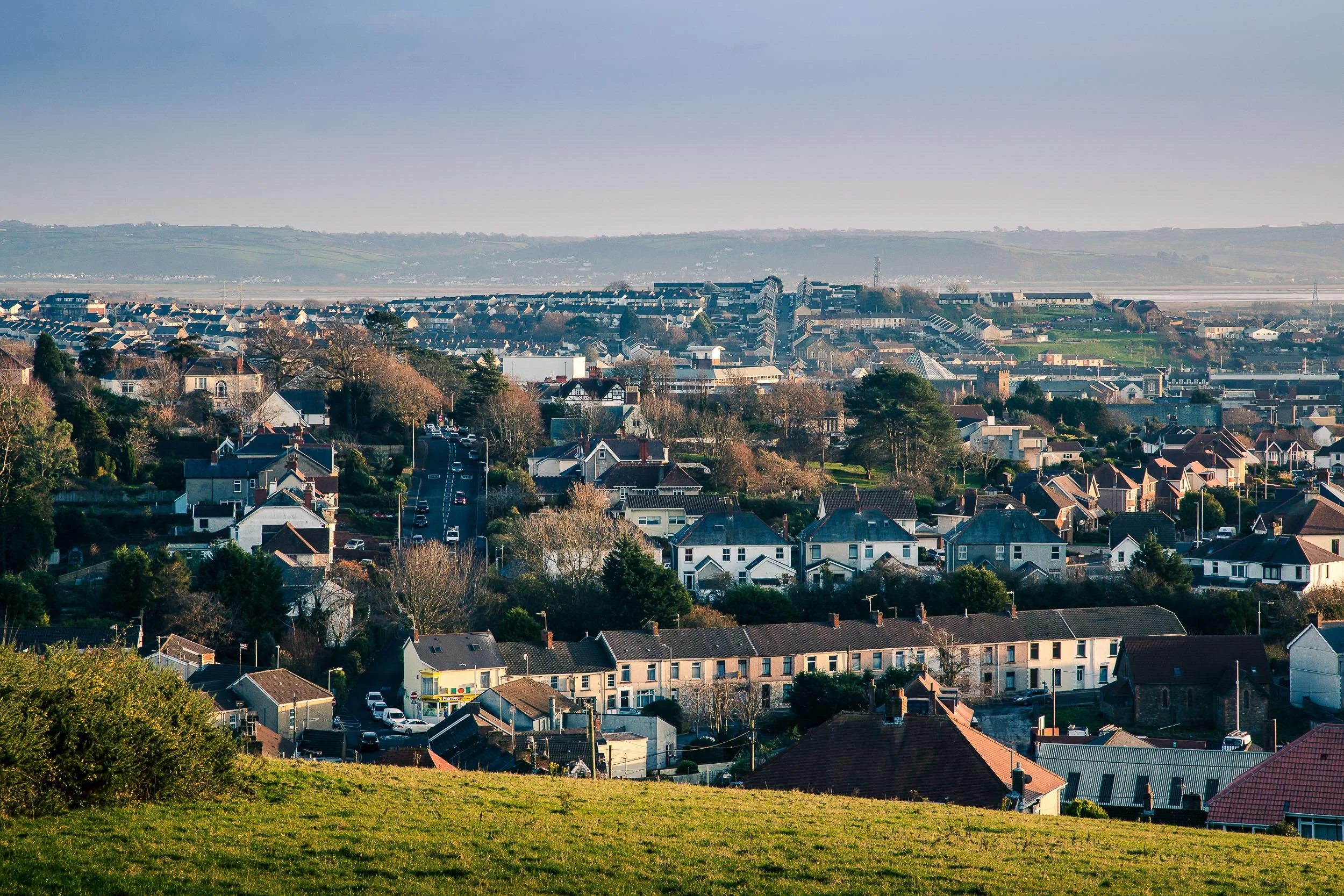 A panoramic view of a city with residential houses, trees, and buildings, with rolling hills in the background during daytime.