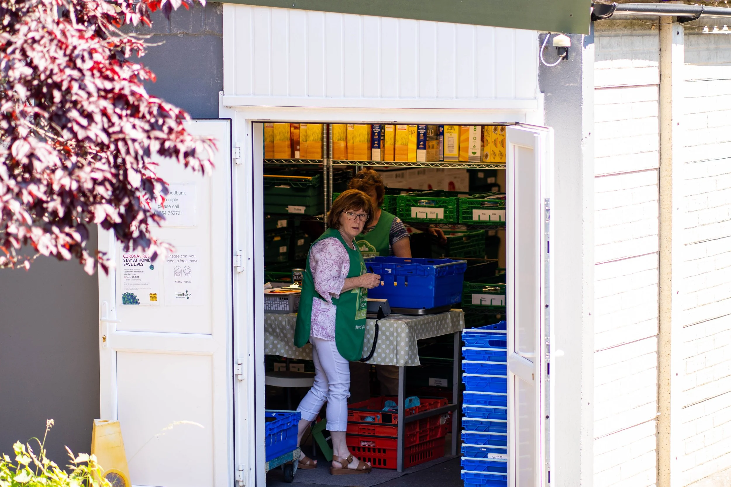 Women working in a small grocery store or market kiosk with shelves of boxed products behind them. One woman is looking out of the open door, wearing glasses and a green apron.