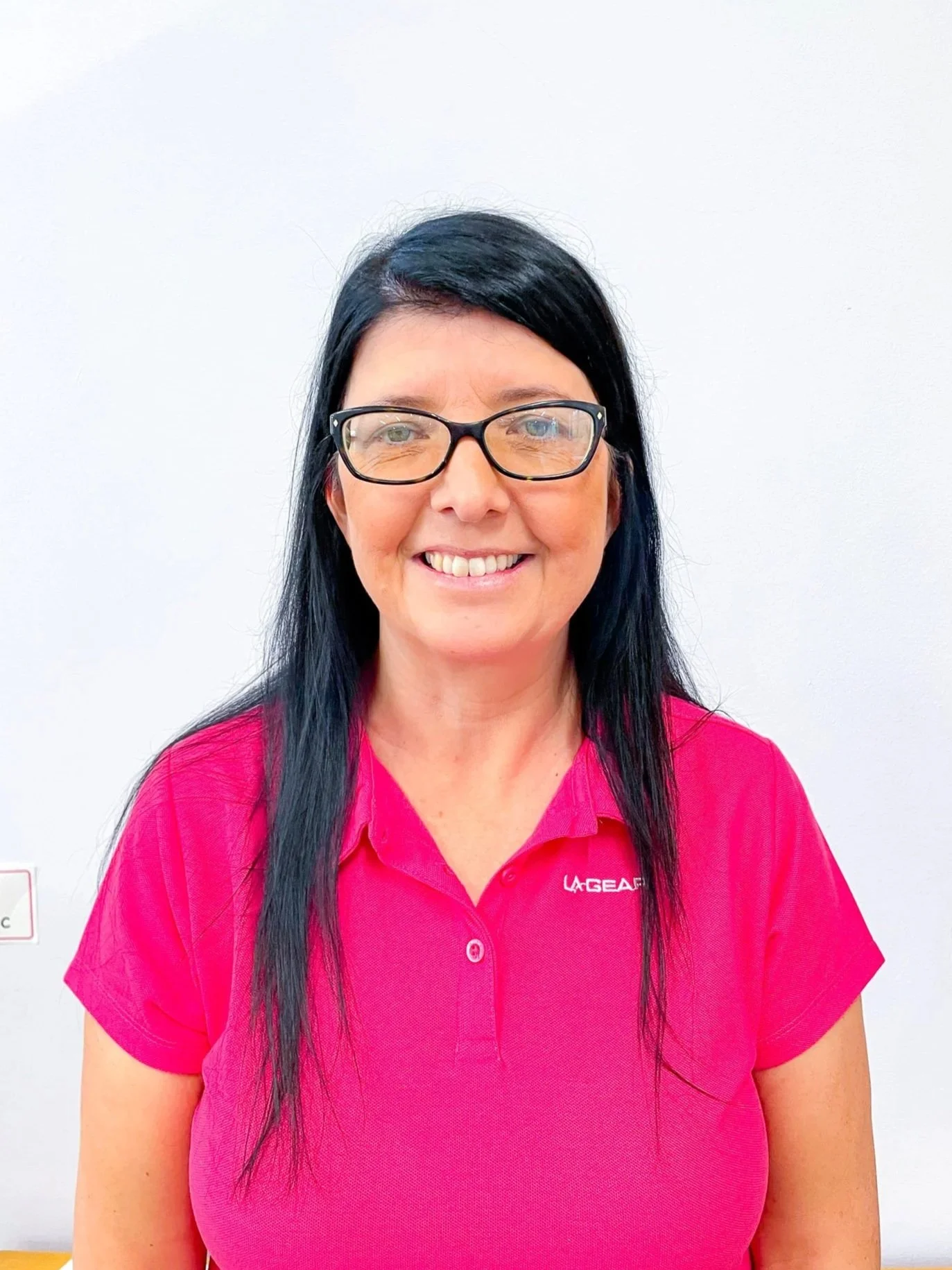 A woman with long brown hair and blue eyes smiling slightly, standing outdoors in front of a windowed building.