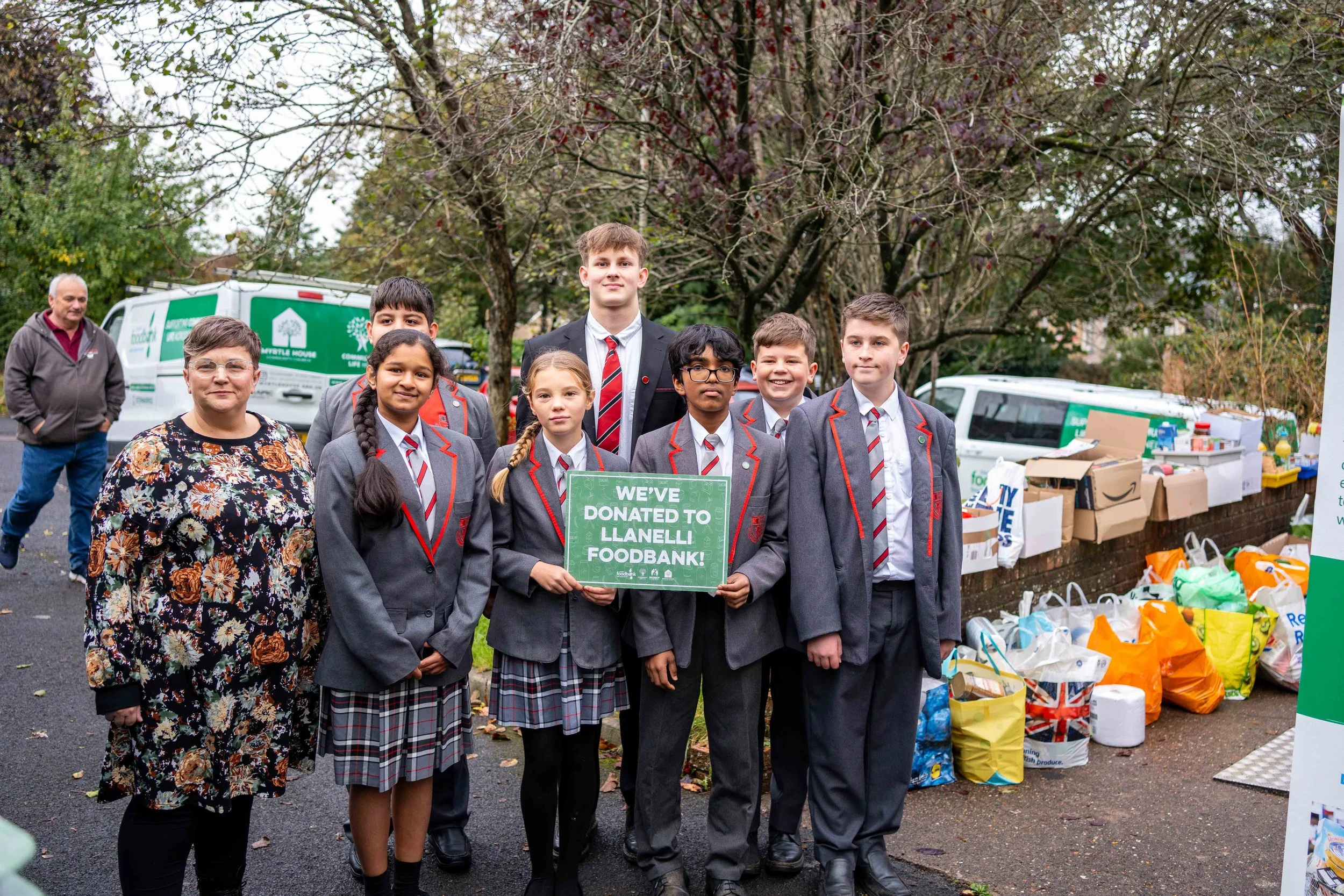 Group of schoolchildren and adult standing in front of food donation collection table, one girl holding a sign that reads 'We've donated to Llanelli Foodbank', outdoors in autumn.