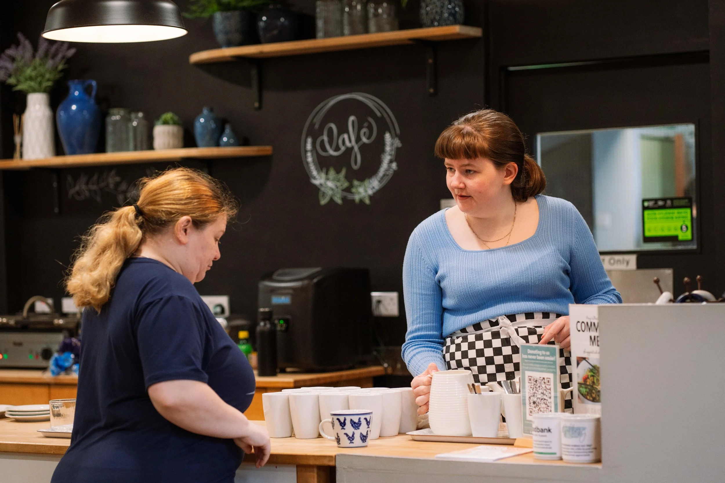 Two women conversing at a coffee shop counter, with one preparing drink and the other placing an order, in a cozy cafe setting with decorative vases on shelves.