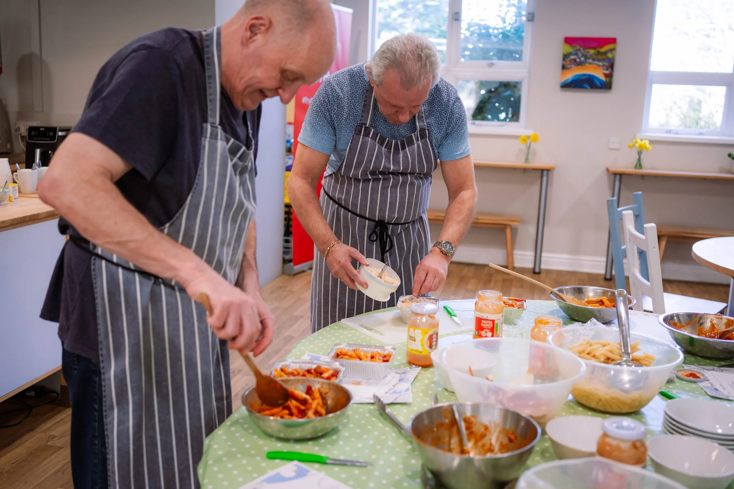 Two elderly men wearing striped aprons prepare food together in a bright kitchen, with bowls of pasta, sauce, and ingredients on the table.