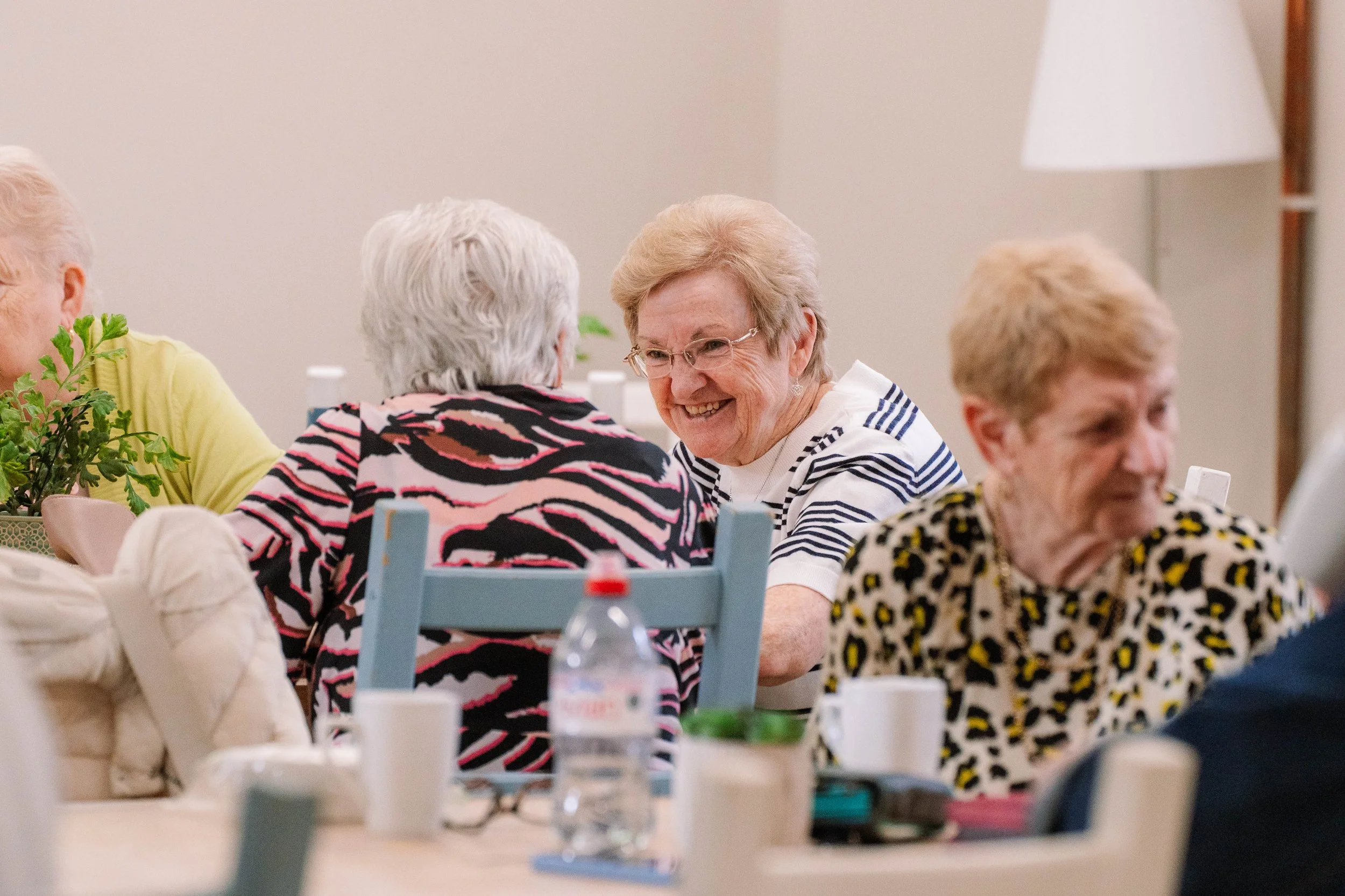 Group of elderly women sitting around a table, engaged in conversation, smiling, with cups, a water bottle, and plants on the table.