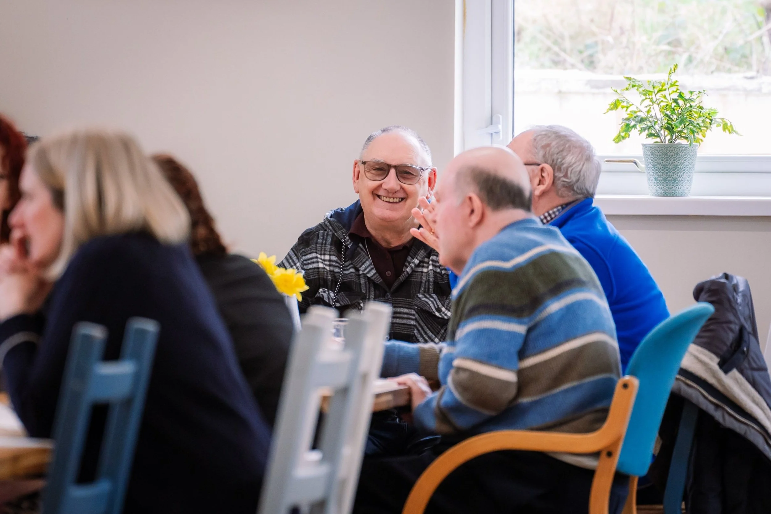 A group of elderly people sitting around a table, enjoying a social gathering indoors, with one man smiling and another talking.
