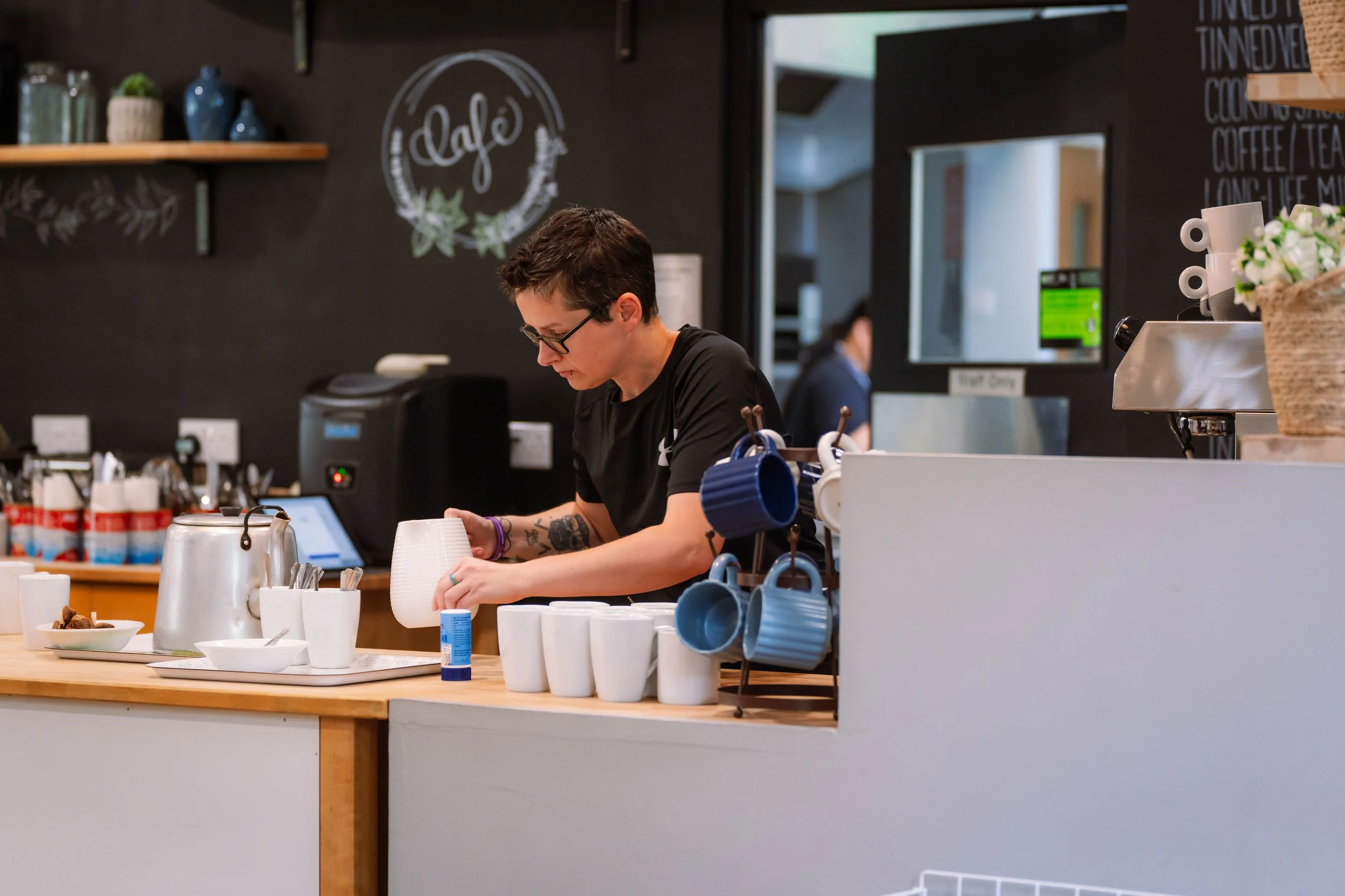 Barista preparing drinks at a coffee shop counter with cups, mugs, and coffee supplies visible.