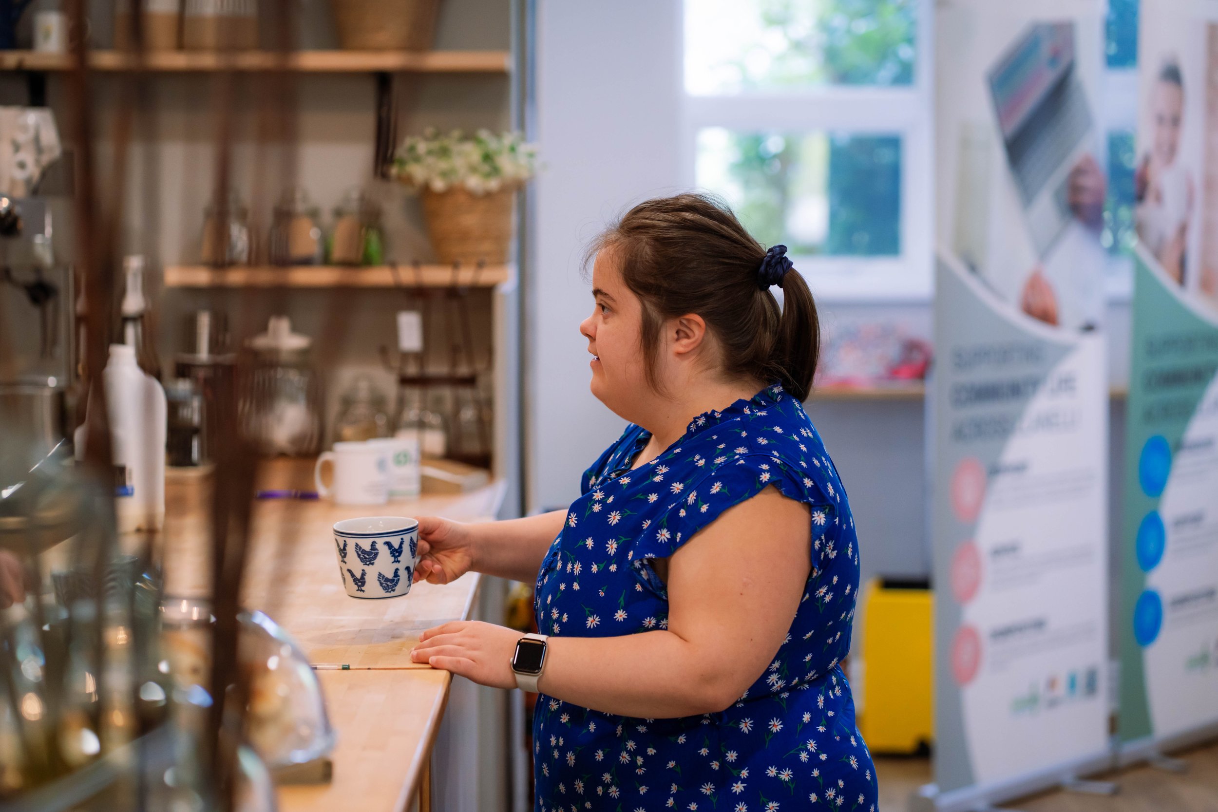 A woman with a ponytail and wearing a blue dress with daisy pattern is holding a blue and white mug at a coffee shop counter.