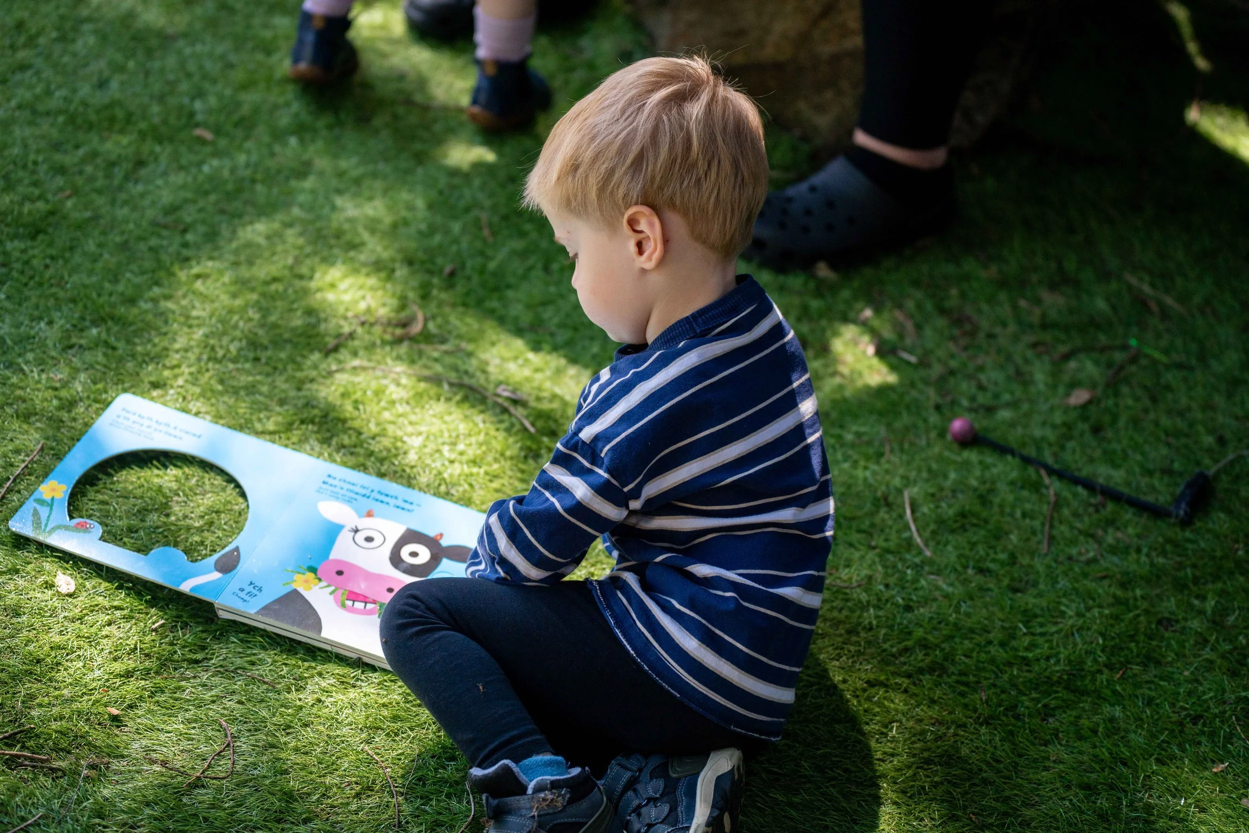 A young boy sitting on the grass reading a children's book with a cartoon cow on the page, outdoors in sunlight with play equipment nearby.