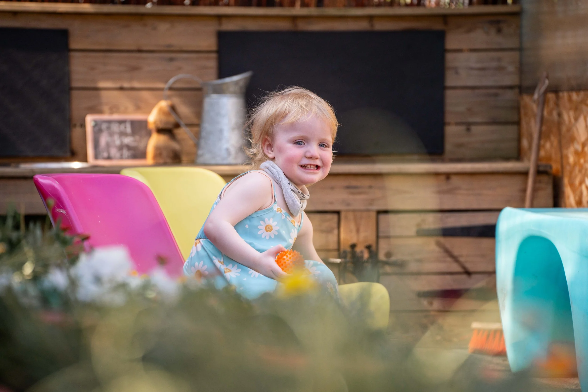 A young child with blonde hair smiling while sitting on a colorful yellow chair in an outdoor setting with wooden walls, surrounded by gardening tools and decorations.