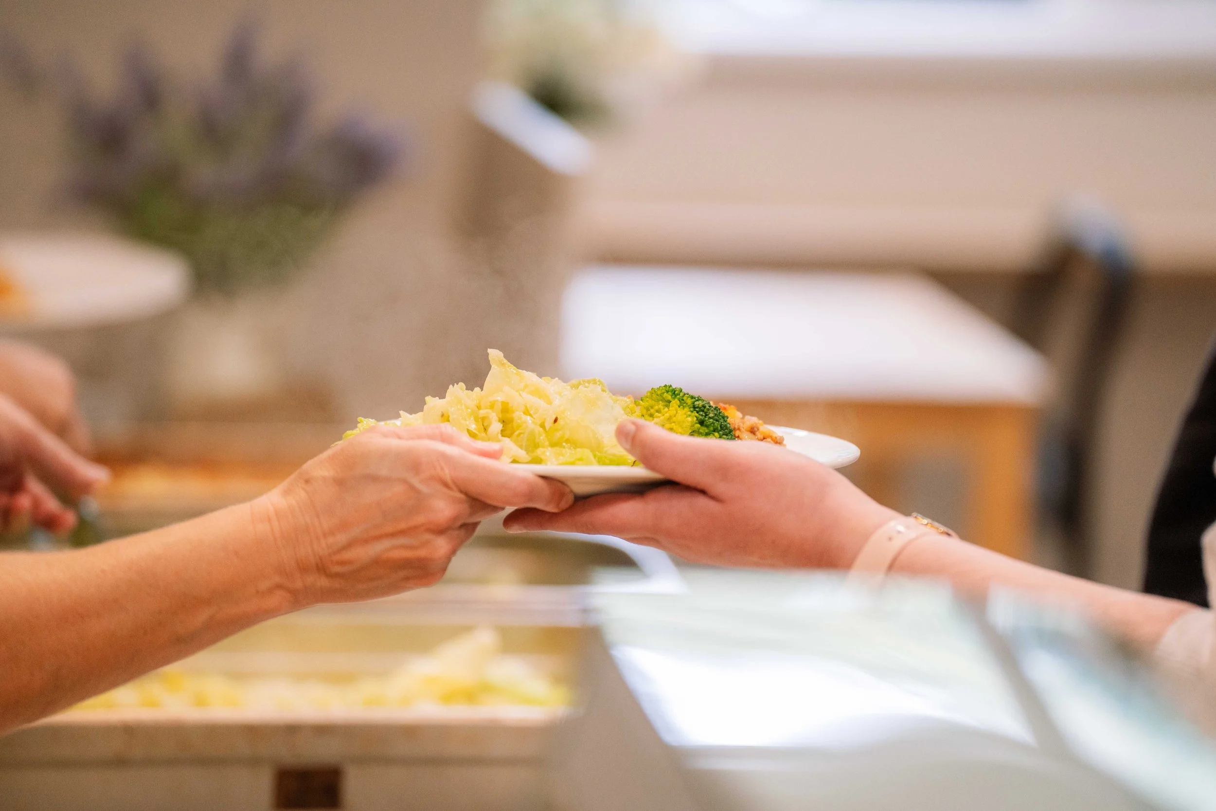 A person is receiving a plate of food, which includes mashed potatoes and broccoli, from another person at a serving area.