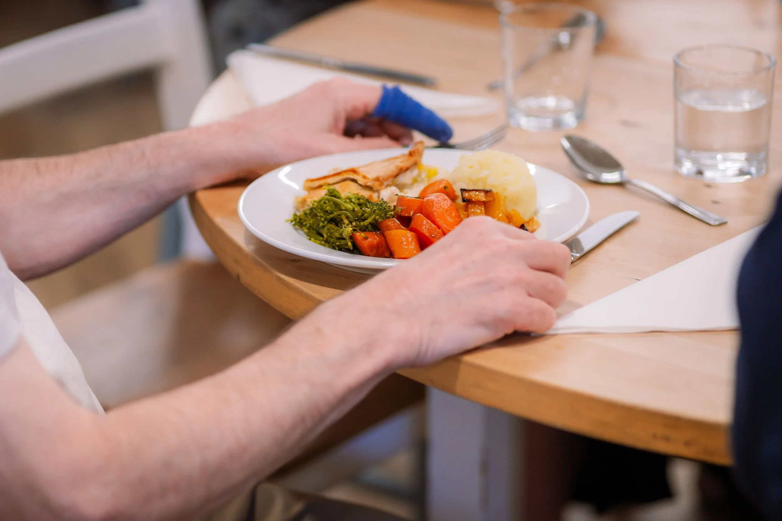 Person with a blue glove holding a plate of food at a dining table with water glasses, a fork, a spoon, and a napkin.