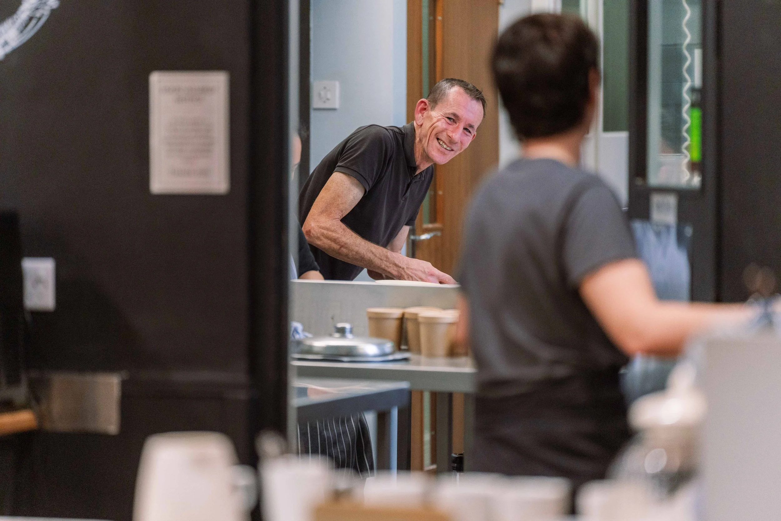 A man in a black shirt smiling and leaning over a counter, viewed through a door frame, as he interacts with a woman working in a kitchen or cafeteria.