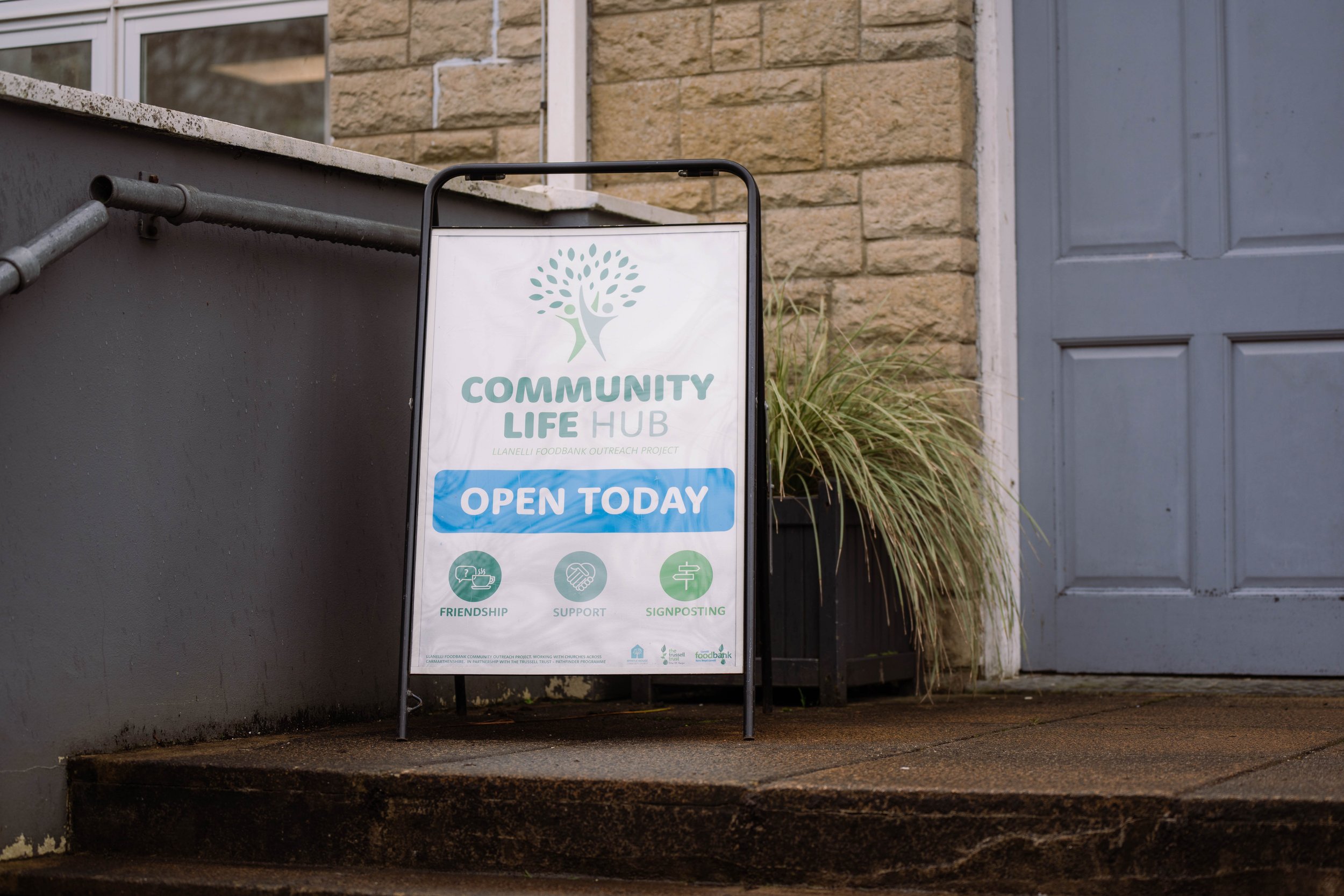 Signboard outside a building promoting the Community Life Hub, indicating it is open today with icons representing friendship, support, and signposting.