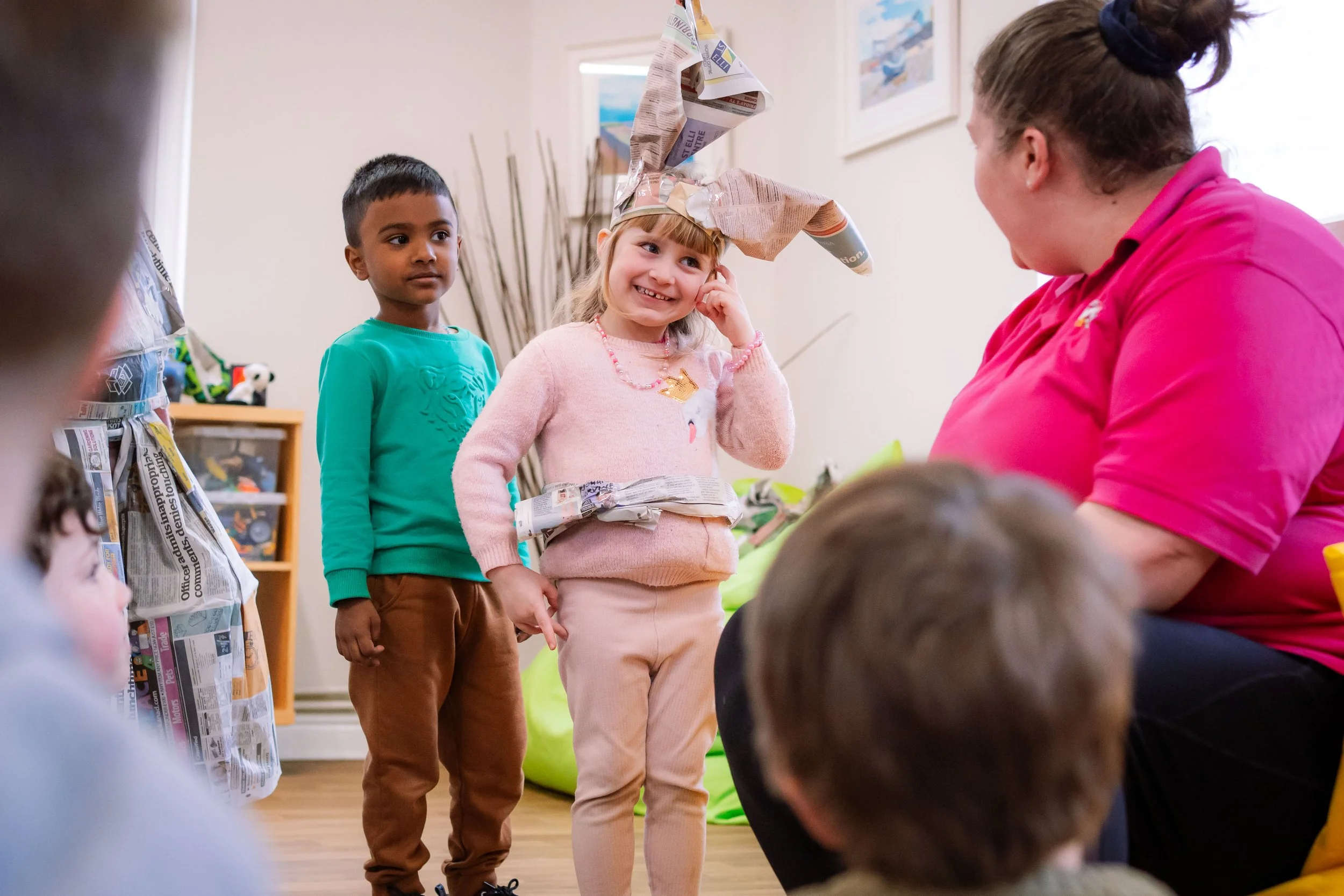 A girl with a paper hat on her head is smiling and touching her face, standing next to a woman in a pink shirt. A boy wearing a turquoise shirt and brown pants stands nearby, with other children partially visible. The setting appears to be a room with artwork on the walls and a gift bag made of newspaper.