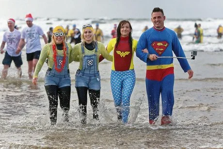 Group of four people dressed as superheroes walking in the ocean waves at the beach, smiling and enjoying the water.