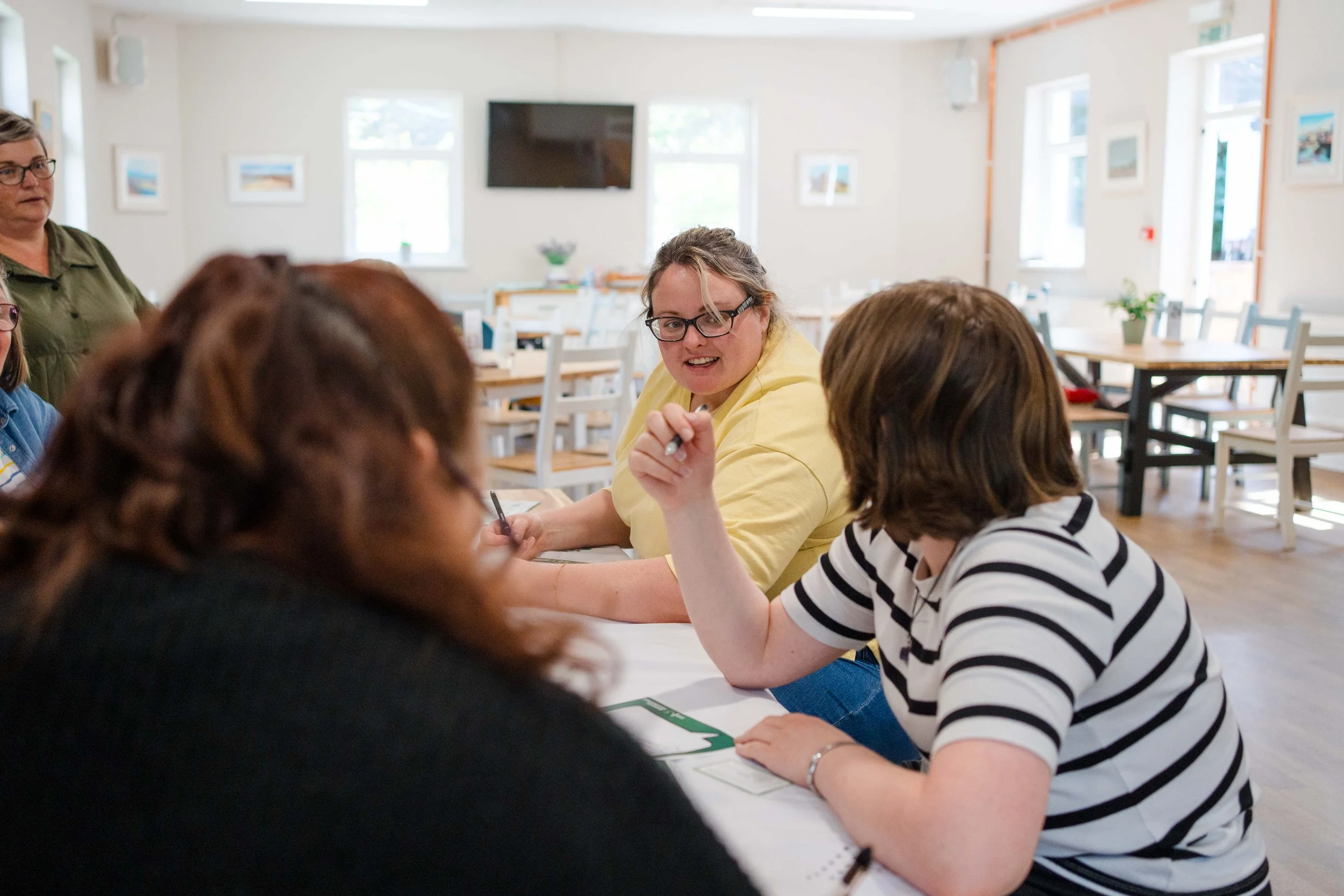 A group of diverse women sitting at a table in a bright, casual meeting room, engaged in conversation. One woman in a yellow shirt is speaking and gesturing, while others are listening. The room has large windows, framed pictures on the walls, and a television.