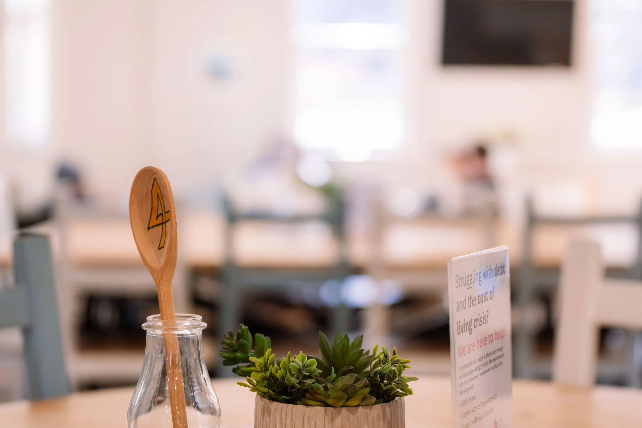 A table decorated with a small potted succulent plant and a clear glass bottle with a wooden spoon inside it, in a bright and blurred indoor setting.