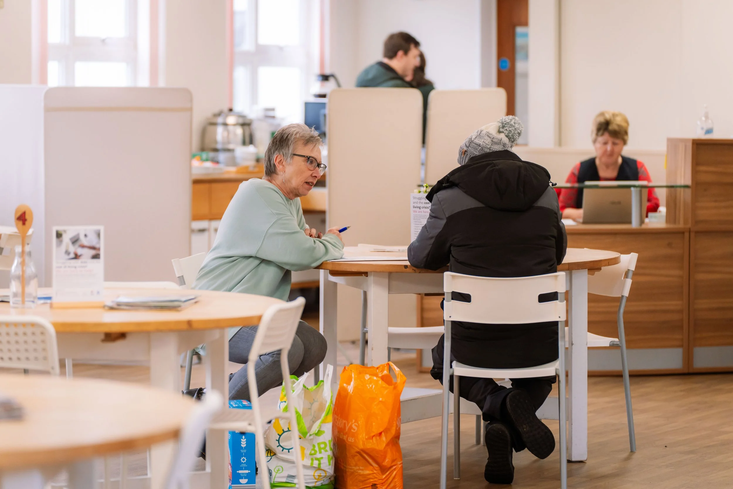 Two women sit at a round table engaged in conversation in a cafe or community center, with one holding a pen. Behind them are people working at a counter, with windows letting in natural light. Shopping bags are placed on the floor beside one of the women.