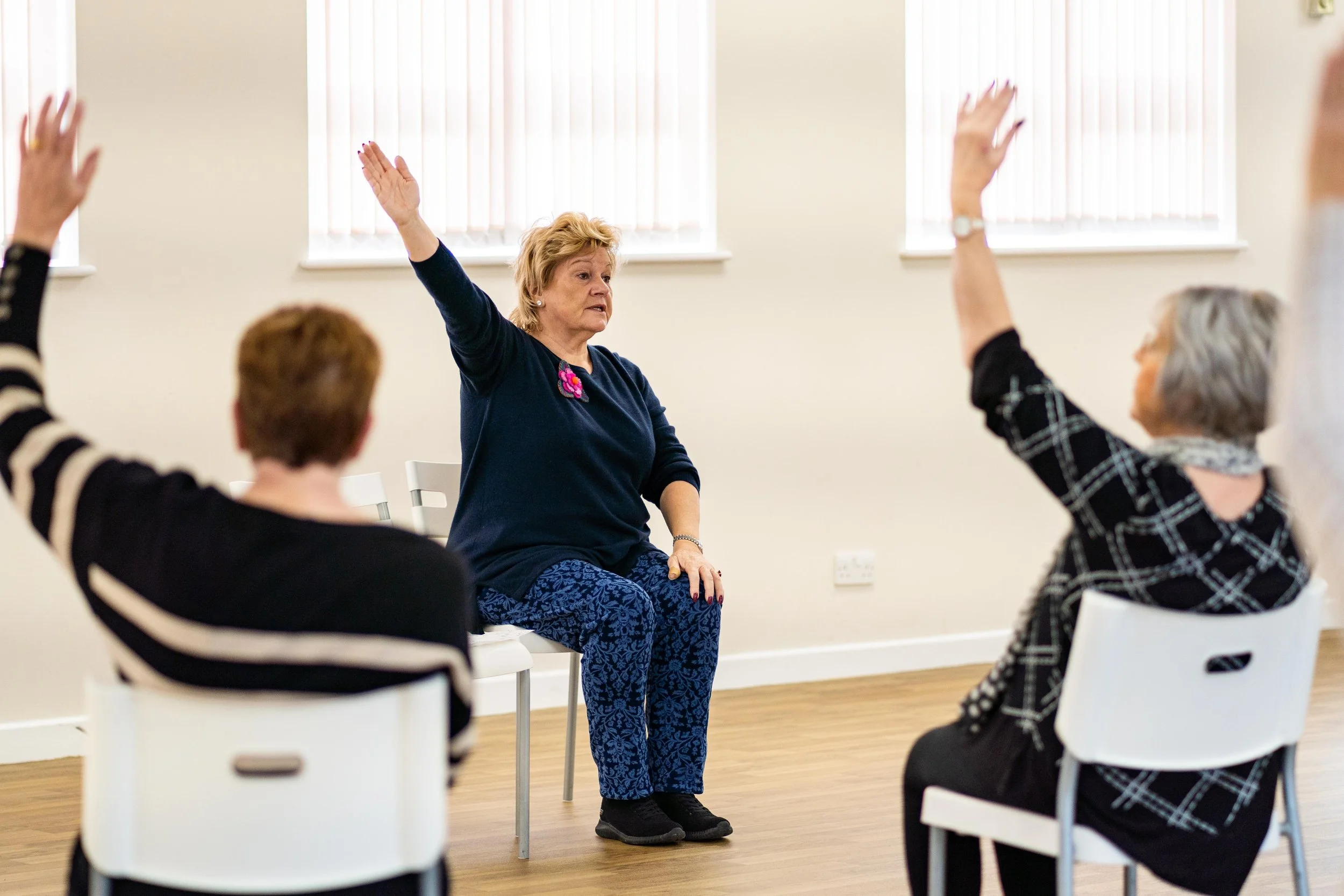 A group of older women participating in a seated exercise or wellness class in a bright room, with the instructor guiding them.