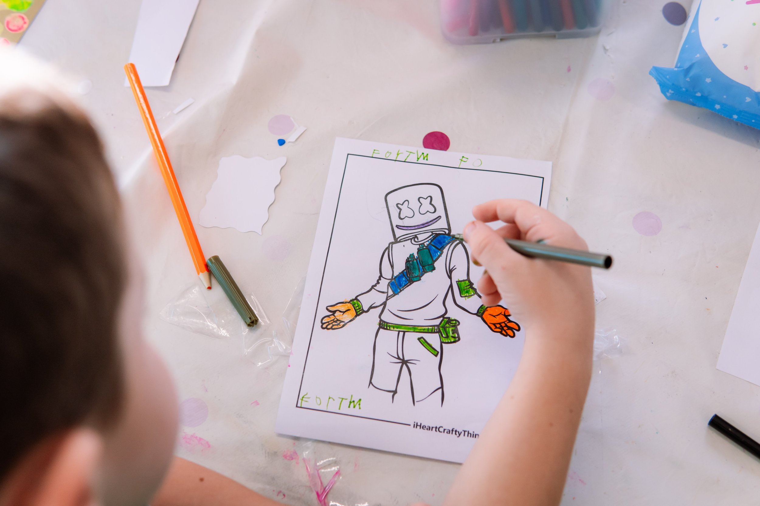 A child coloring a drawing of a robot on a white sheet of paper, sitting at a table surrounded by coloring supplies and confetti.