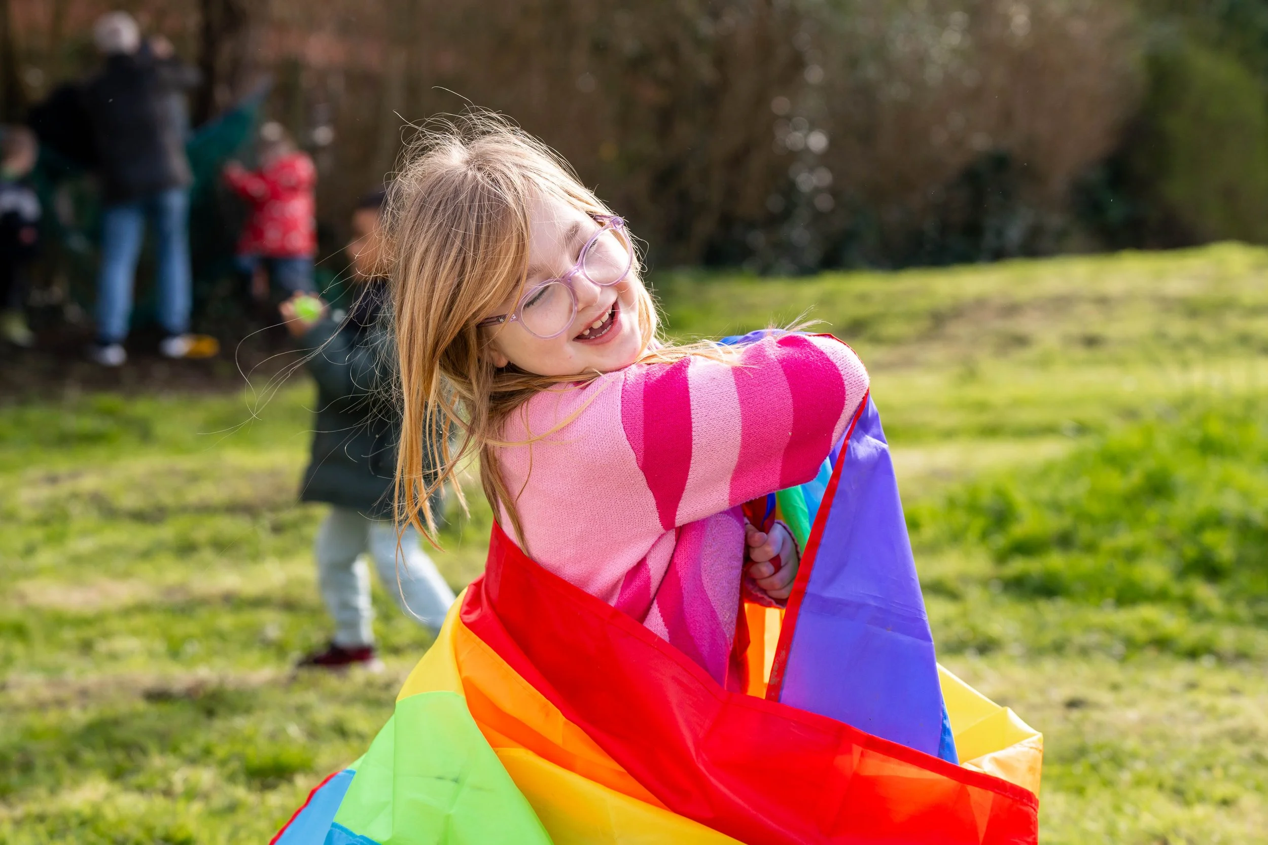 A young girl with glasses and a pink striped sweater smiling and playing outdoors with a colorful rainbow parachute or kite, with children and adults in the background on a grassy area.
