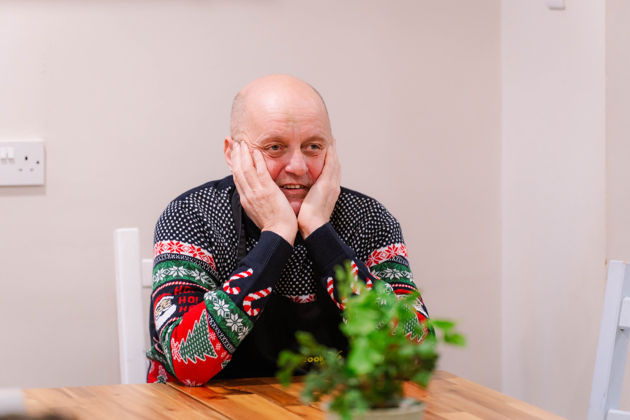 A bald man wearing a festive Christmas sweater sits at a wooden table, resting his face in his hands, with a surprised or overwhelmed expression.
