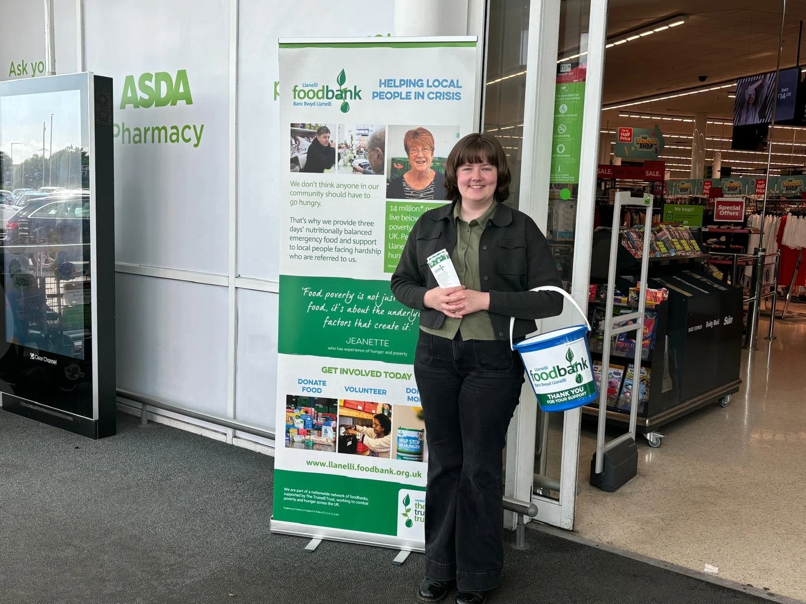A young person standing outside a food bank in a supermarket, holding a donation bucket and pamphlet, smiling. Behind them is a large banner with information about the food bank and its mission.