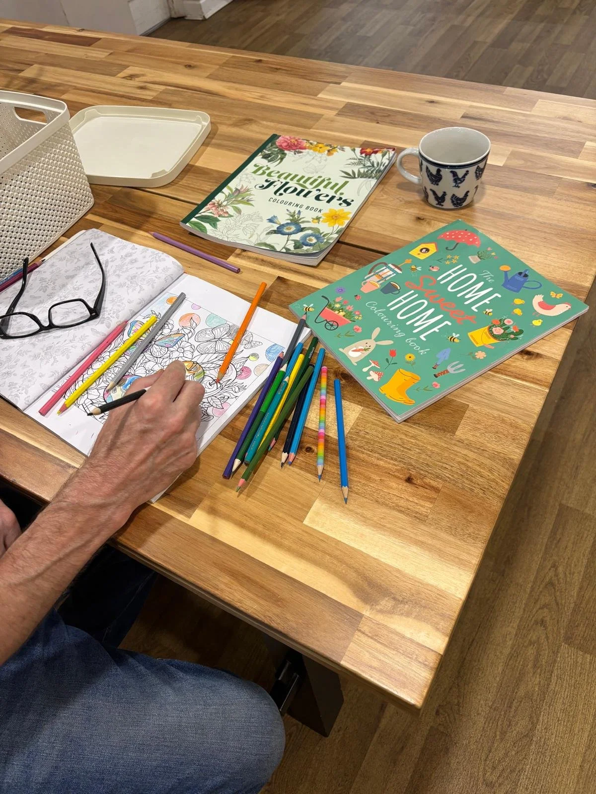 A person coloring in a coloring book at a wooden table surrounded by books, colored pencils, and other drawing supplies.