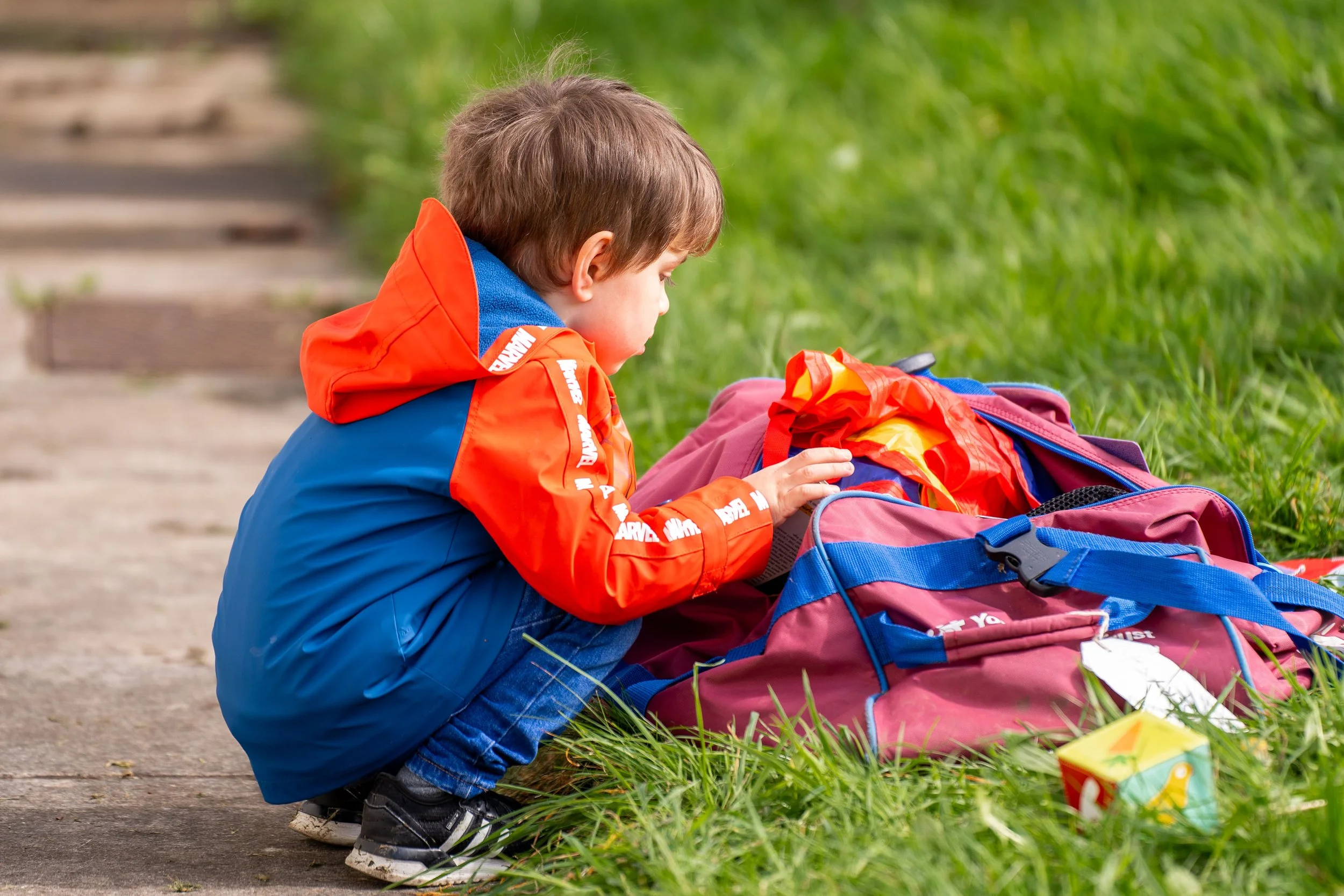 Young boy in a colorful jacket crouching and looking into a backpack on the grass next to a paved path.