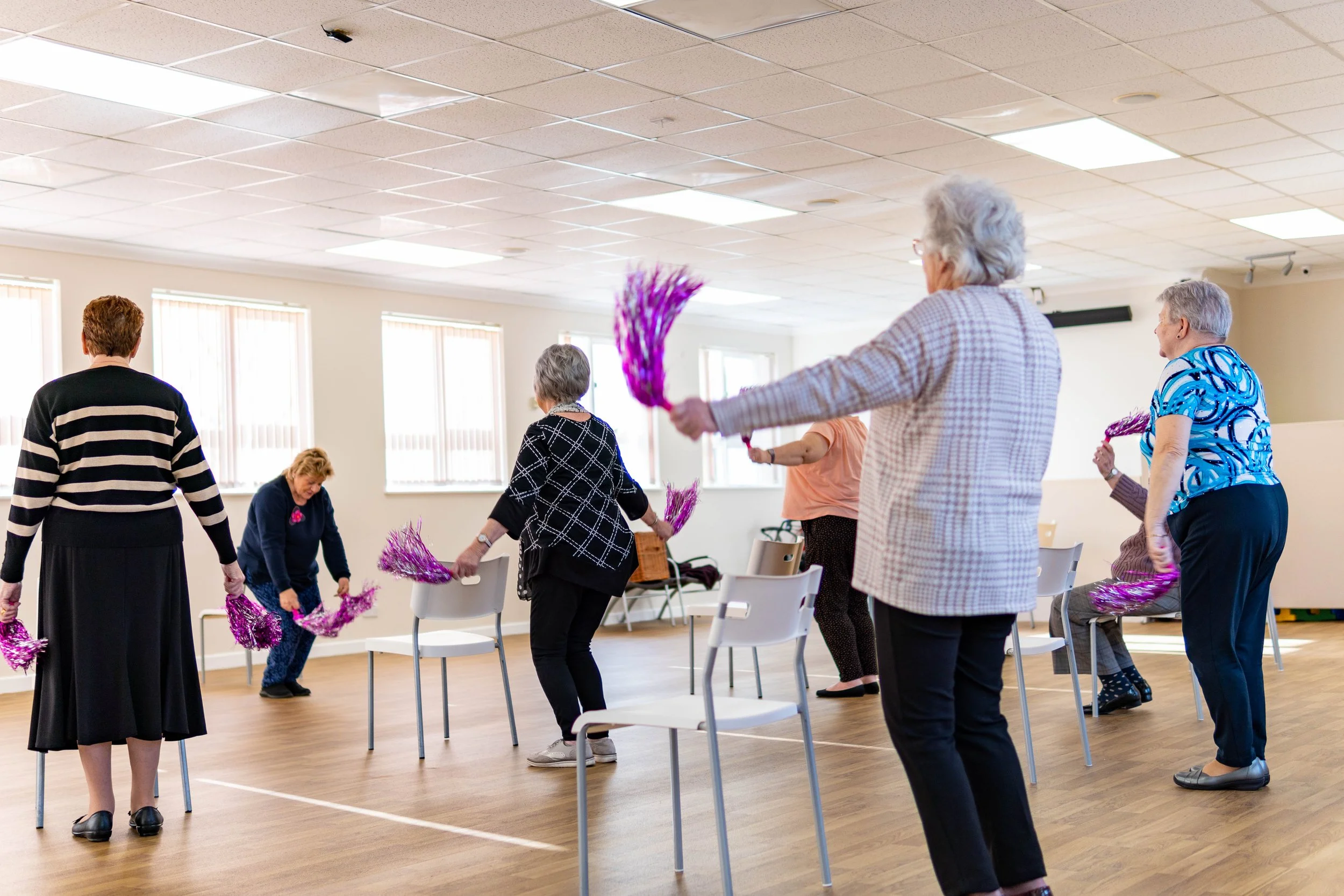 Group of elderly women participating in a seated exercise class in a bright, spacious room, holding purple tinsel pom-poms.