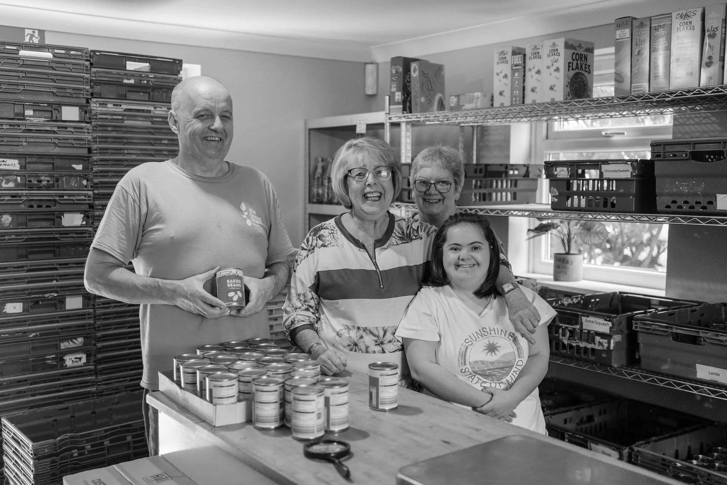 Four people standing inside a food pantry or storage room, with shelves of canned goods and boxed cereal behind them, smiling at the camera.
