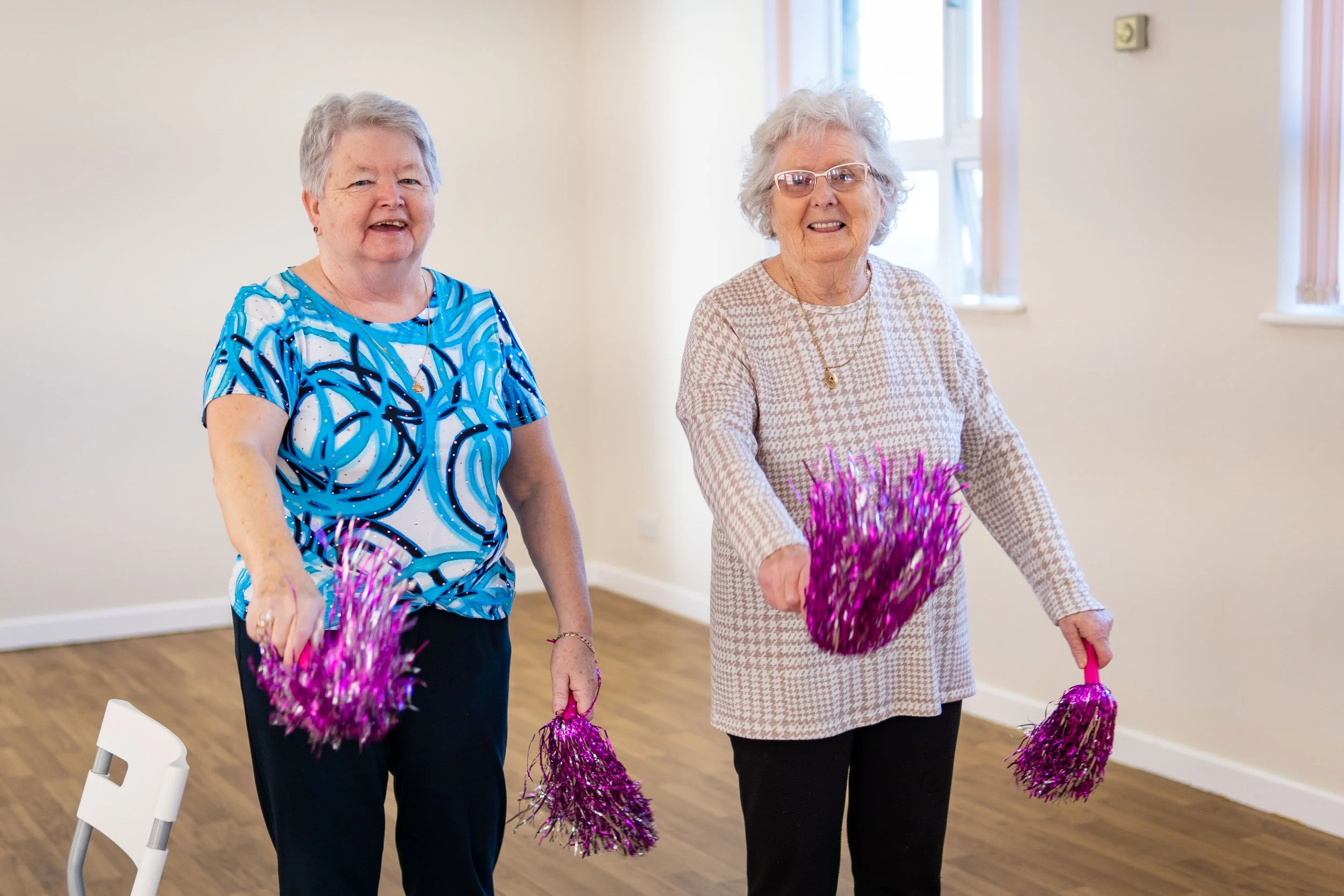 Two elderly women dancing indoors while holding pink tinsel pom-poms.
